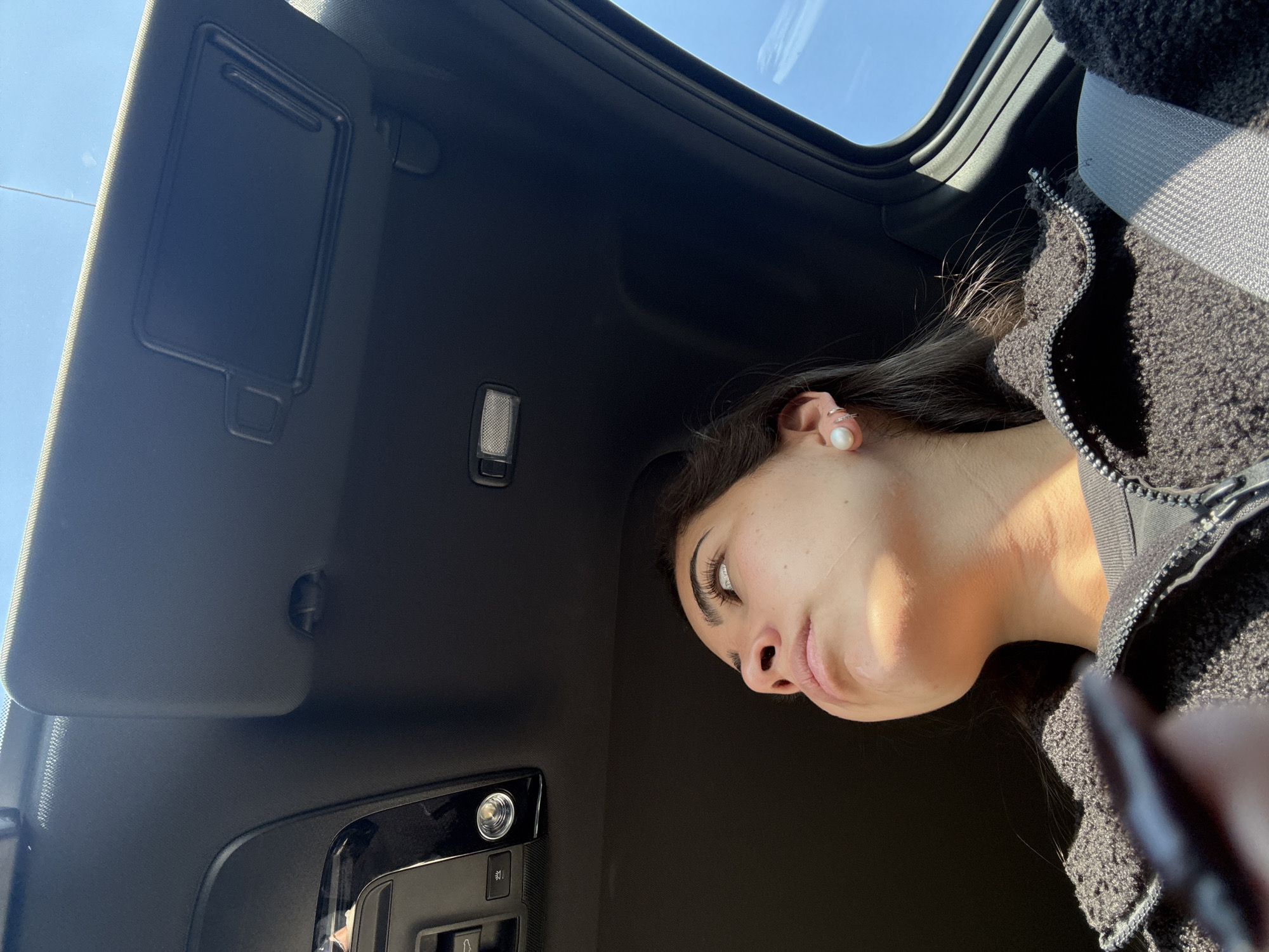 A woman is in a car with an open sunroof. She is wearing a jacket and a pearl earring. The interior of the car is dark. The image was likely taken inside the car.