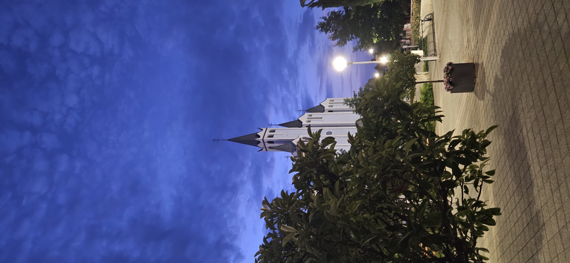 The image depicts a view of the Church of the Holy Spirit in Hévíz, Hungary, at dusk. The sky is a deep, stormy blue with dramatic clouds. The white church, with its distinct black spires topped with crosses, is a prominent feature in the background. In the foreground, a large, leafy tree partially obscures the view of the church and the surrounding area. The ground is paved with square tiles, and a modern streetlamp casts a warm glow. Further back, there are more trees, a bench, and a planter with pink flowers. The scene conveys a sense of quiet contemplation as daylight fades.