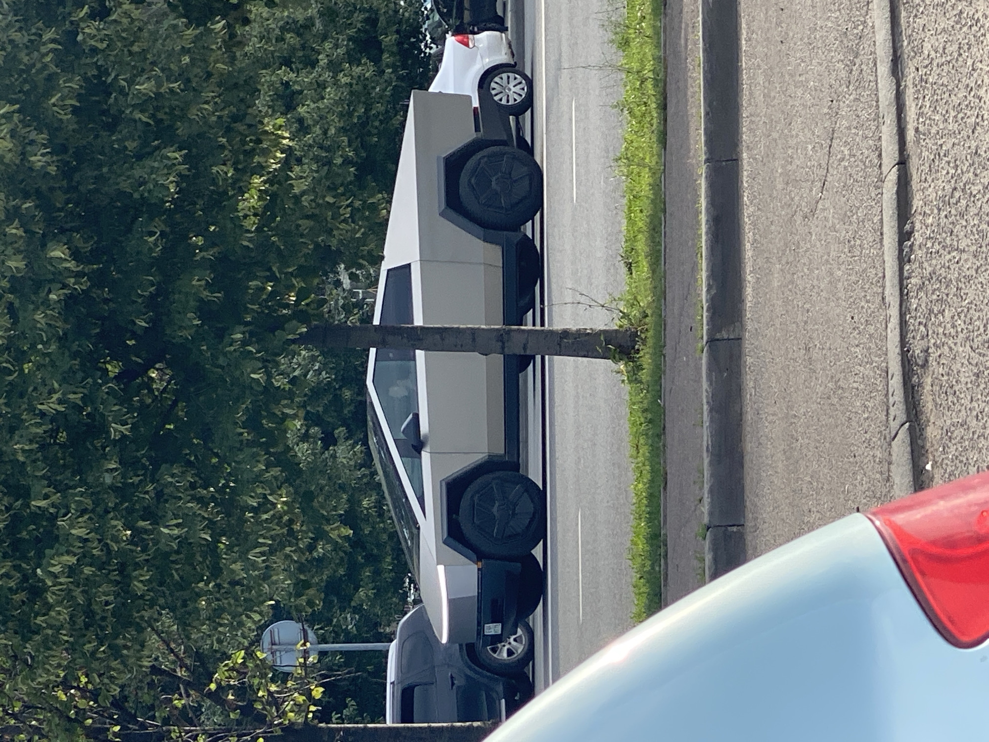 A Tesla Cybertruck is visible from the side, driving on a paved road. The truck is a light gray color and has a distinctive angular design. It is partially obscured by trees and the foreground of another vehicle. In the background, other cars, including a white sedan, are also on the road. The scene appears to be an urban or suburban environment, with a grassy median strip and trees lining the road. The light suggests it is daytime, possibly late afternoon. The specific location is the city of Kazan, Russia.