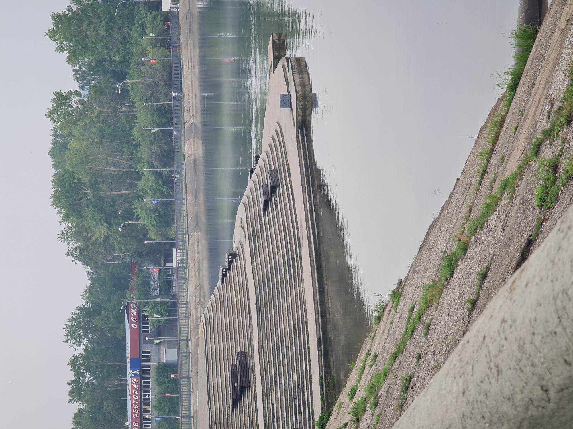 The image captures a wide view of a waterfront area in Unknown, Unknown, featuring a prominent concrete amphitheater-like staircase descending towards the water. The amphitheater has several levels with dark wooden benches strategically placed on some of the steps. In the background, a building with a red roof and the word "РЕСТОРАН" (Restaurant) in Cyrillic is visible, along with a line of trees that forms the horizon. A metal railing separates the park area from the water's edge. The water itself is calm, reflecting the overcast sky and the surrounding greenery. The foreground shows a sloped concrete embankment with patches of green grass growing through it. The overall atmosphere is serene and somewhat muted due to the cloudy weather. There are no visible people in the immediate foreground or on the amphitheater steps, but a few figures can be seen walking along the promenade behind the railing. The scene appears to be a public recreational space.