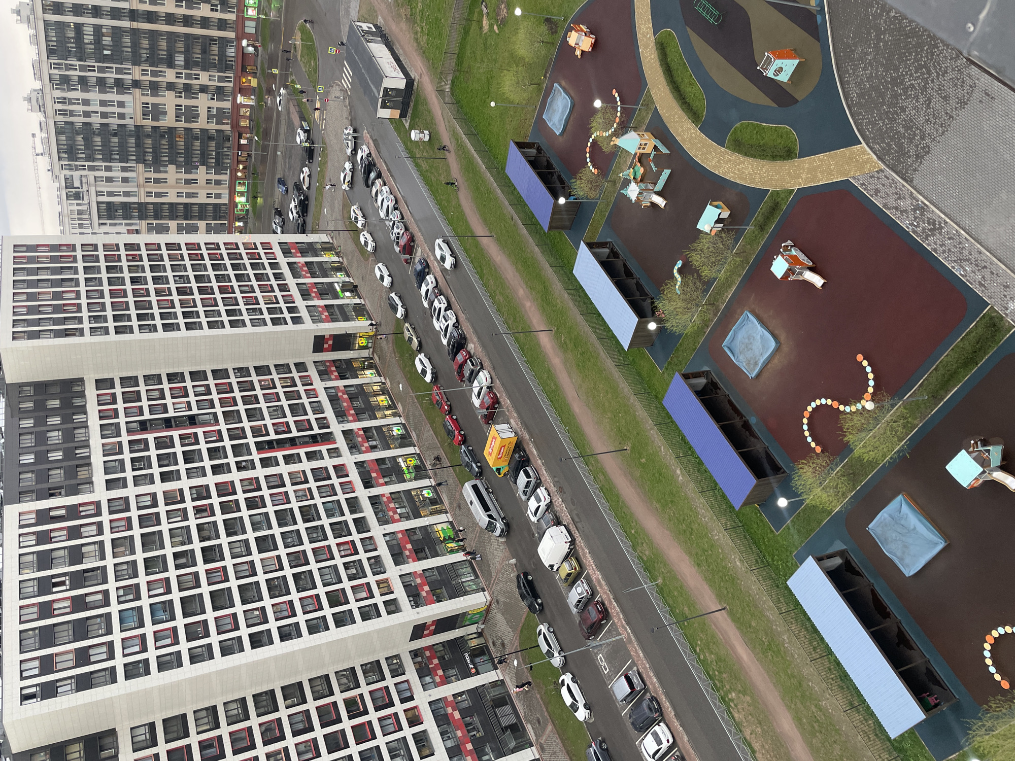 The image shows an aerial view of a residential area in an urban setting, likely in Unknown, Russia. Two tall apartment buildings with a grid-like pattern of windows dominate the left side of the frame. A street runs between the buildings and a playground. Cars are parked along the street. Pedestrians can be seen on the sidewalk. The playground on the right has colorful structures and play equipment. A pedestrian crosswalk is visible near the top of the frame. The overall environment suggests a densely populated, planned residential area.