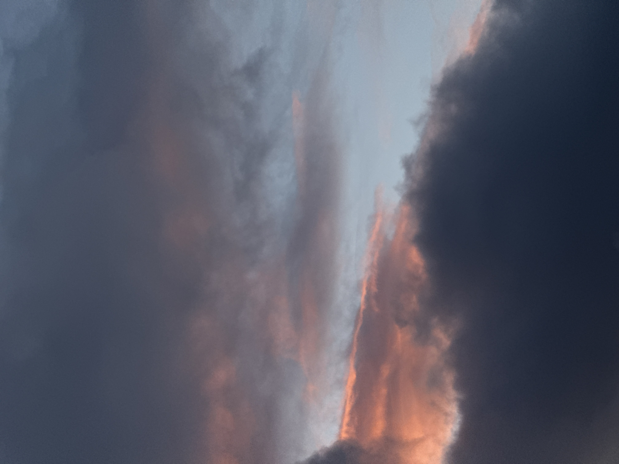 The media file presents a dramatic and atmospheric sky scene, dominated by a dynamic interplay of clouds at what appears to be either sunrise or sunset. The sky is filled with expansive cloud formations, exhibiting a striking range of colors.
Large, dark grey and deep blue clouds occupy significant portions of the frame, suggesting heavy cloud cover or an impending weather event. These darker masses are contrasted vividly by streaks and patches of brilliant orange and soft pink, which illuminate the edges and undersides of some clouds. These warm hues indicate that the sun, though not directly visible, is low on the horizon, casting its light onto the cloud formations. Lighter, more ethereal blue-grey sections of the sky are also visible between the darker cloud masses.
There are no people, man-made objects, or specific activities depicted in the image. The scene is purely a natural landscape of the sky, focusing on the atmospheric conditions and the captivating effect of light on clouds. No visible text is present. The general impression conveyed is one of awe-inspiring natural beauty and dramatic weather dynamics, consistent with a spectacular start or end to the day over Fécamp, France.