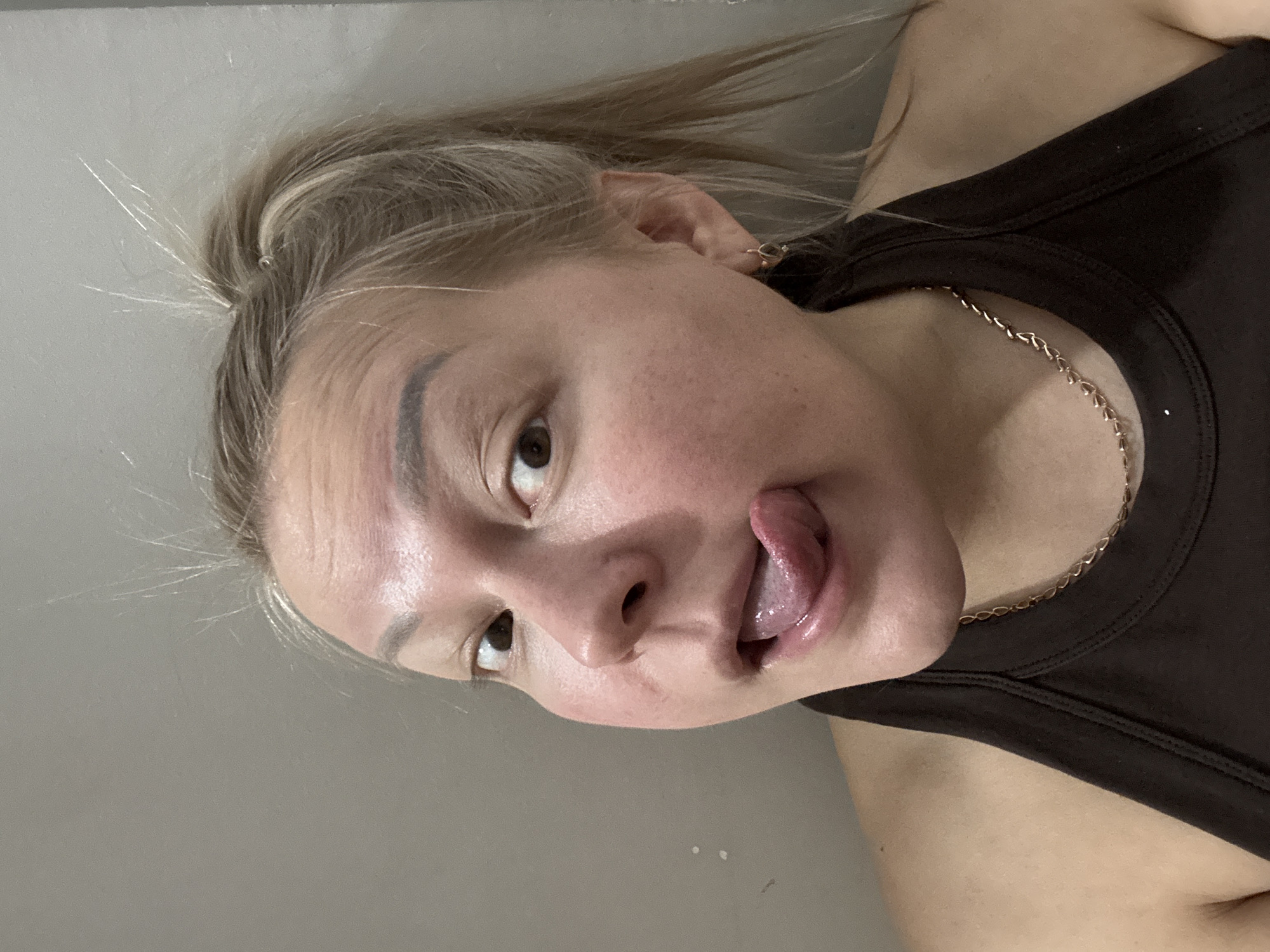 A close-up, slightly off-kilter, indoor photograph captures a person with fair skin and light brown or blonde hair. The hair is pulled back, revealing the forehead and a portion of the left ear. The person is sticking their tongue out and looking slightly to their left. They have brown eyes, visible eyebrows, and a light complexion.
The person is wearing a dark-colored tank top, and a delicate gold chain necklace is visible around their neck.
The background is a plain gray wall, providing a neutral backdrop that does not reveal the city of Unknown, Unknown. No other people, subjects, objects, actions, interactions, or environmental details are discernible in the scene.