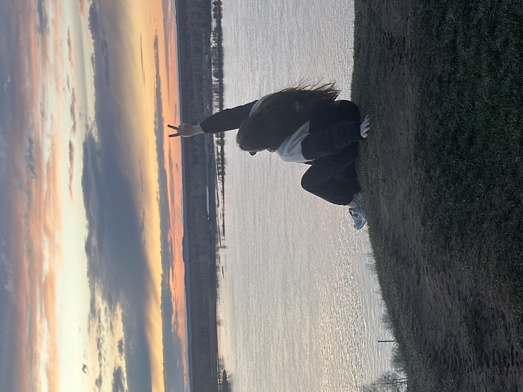 The image captures a person silhouetted against a dramatic sunset over a wide body of water in Ольховка, Russia. The individual is positioned on a grassy and muddy bank in the foreground, facing towards the water and the setting sun. They are in a crouched or squatting posture, with long, dark hair partially obscuring their face. One arm is extended, forming a "peace" sign with two fingers. They are wearing a dark jacket over a light-colored hoodie, dark pants, and light-toned sneakers.
The midground features a calm body of water, likely a river or large lake, reflecting the light from the sky. Across the water, a distant shoreline with faint trees or landmasses is visible. The background is dominated by a vibrant sunset sky, showcasing a blend of warm orange, yellow, and pink hues transitioning into cooler blues and greys, with distinct cloud formations. The overall impression is one of tranquility and contemplation at dusk. No visible text is present in the image.