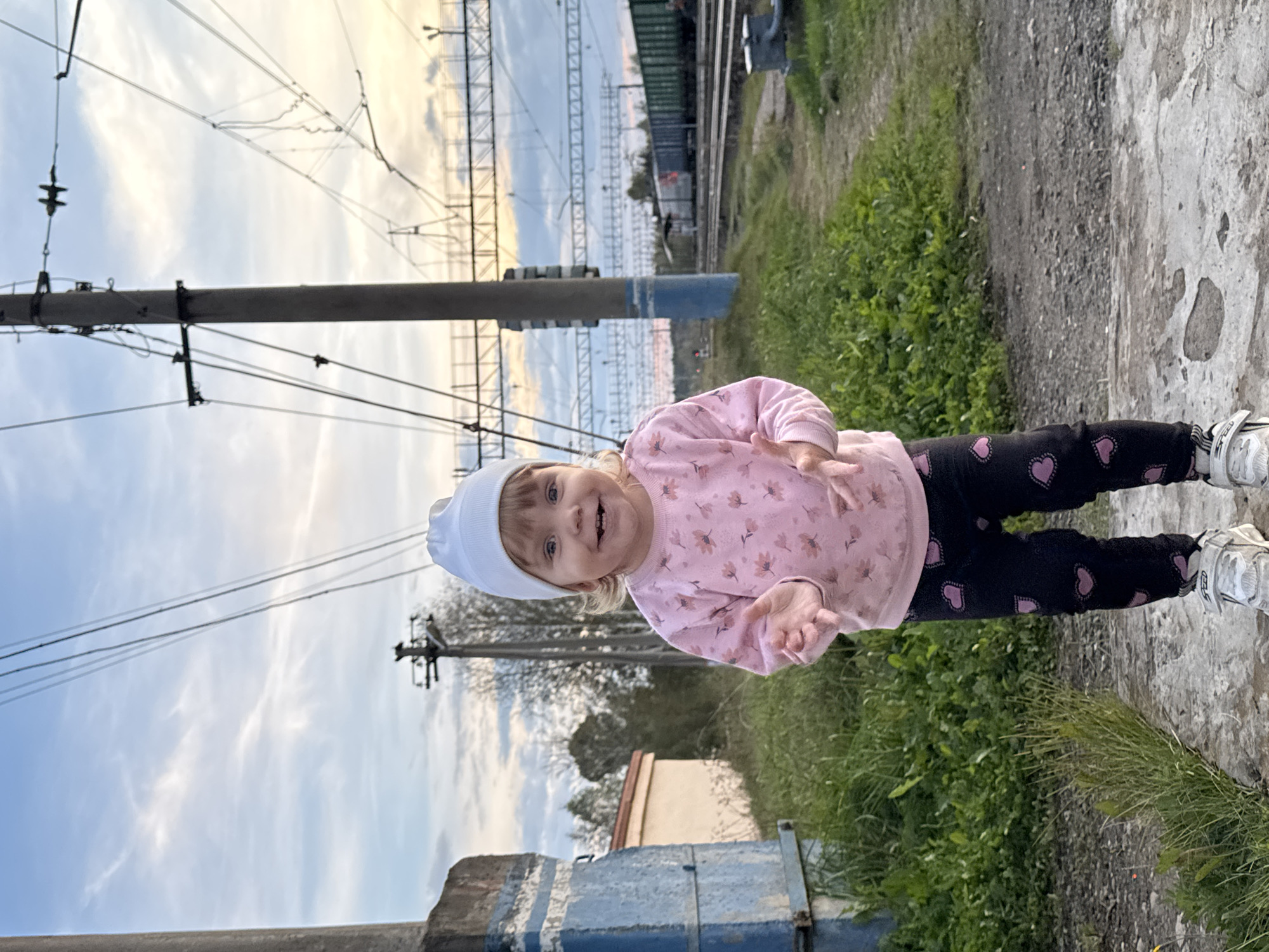 A young girl wearing a pink floral long-sleeved shirt, black leggings with pink hearts, a white beanie, and white sneakers stands on a concrete path. She is facing the camera with her hands extended forward, palms up. Behind her, a train track with overhead power lines runs horizontally across the image. In the background, a train with green and grey carriages is visible. The scene appears to be an outdoor location near a railway in Комарихинский, Russia, with vegetation and a concrete structure to the left. The sky is partly cloudy, with hints of sunset colors.