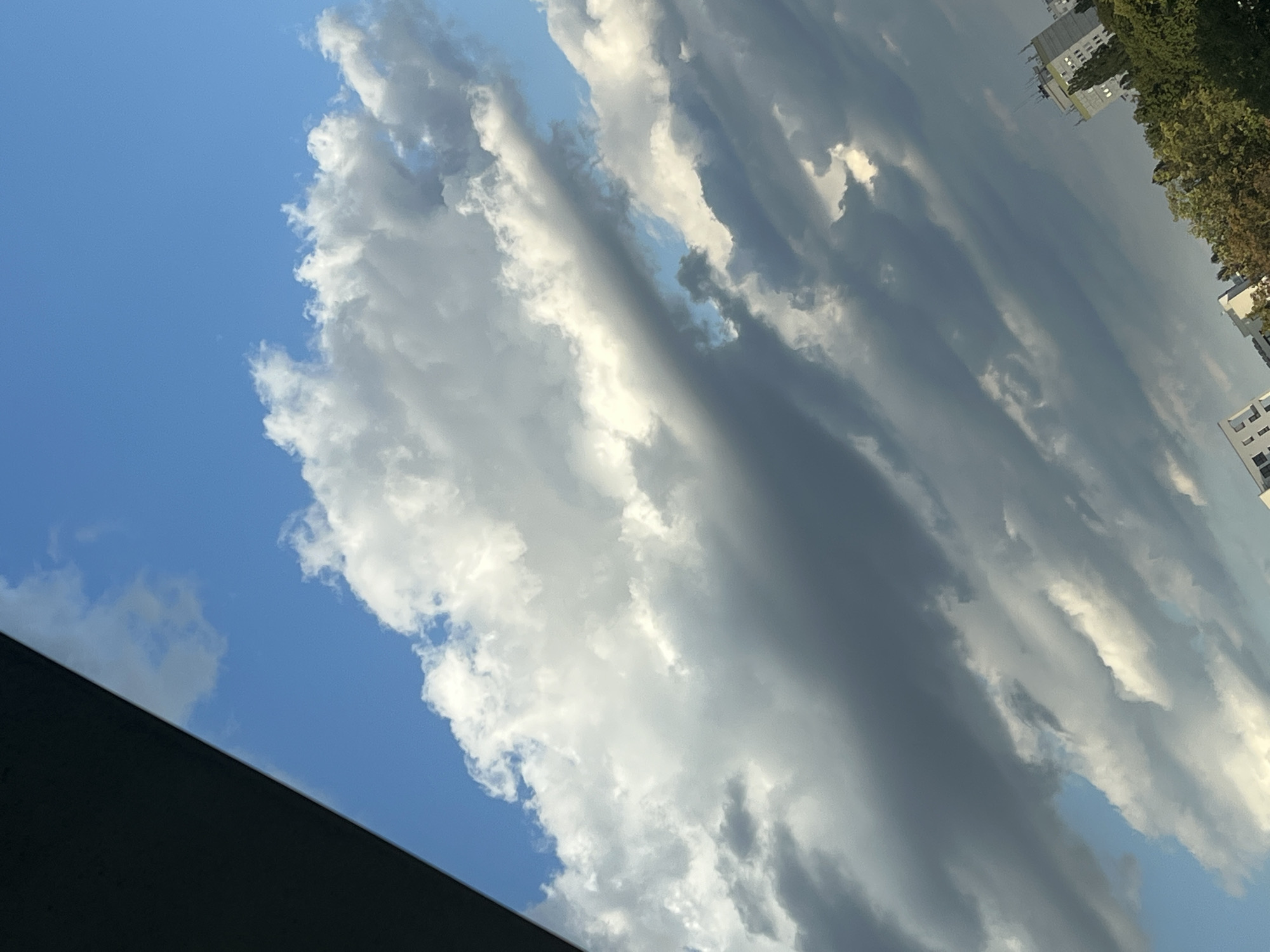 The image depicts a dramatic sky filled with a mix of white and dark gray clouds, interspersed with patches of bright blue sky. The clouds are layered and appear to be moving, creating a sense of depth and dynamism. In the lower right corner, the tops of several buildings and a dense line of green trees are visible, suggesting an urban or suburban environment. The perspective of the shot is tilted, as if taken from a window or elevated vantage point. The lighting suggests it could be late afternoon or early evening, with the sun casting shadows and highlighting the textures of the clouds. The composition is dominated by the sky, with the city elements serving as a grounding detail at the bottom. Based on the architecture and foliage, the scene could be a part of Warsaw, Poland.