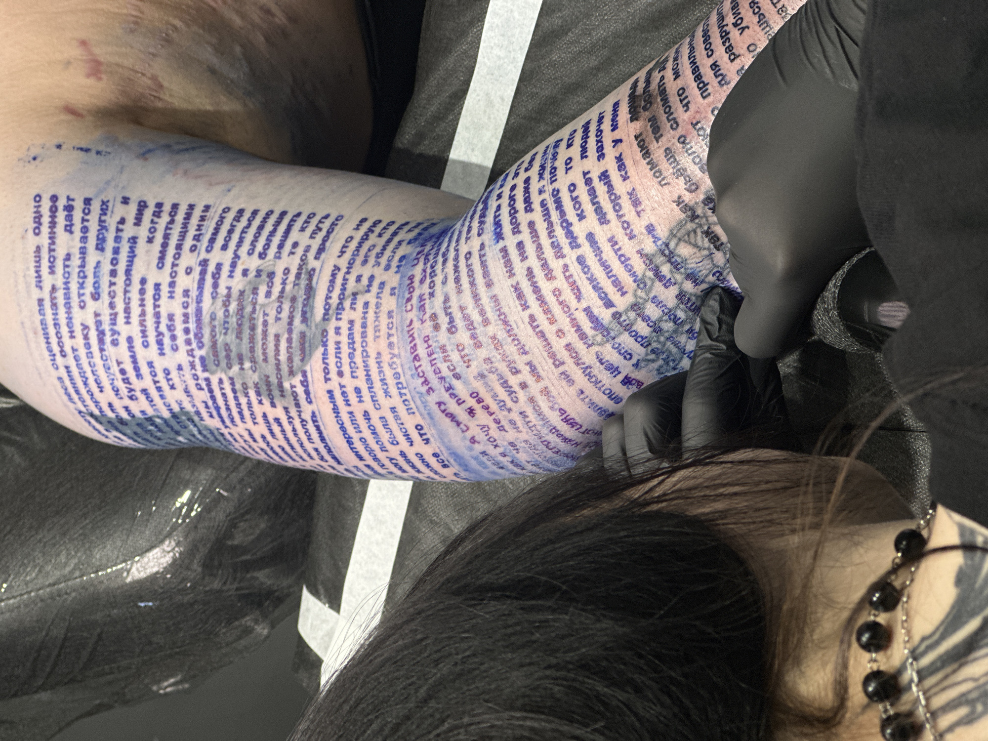 The image shows a tattoo artist in black gloves tattooing Russian text onto a person's arm. The text appears to wrap around the entire forearm. The tattoo artist is visible from the chest up, with dark hair and a necklace. The arm being tattooed rests on what appears to be a glossy black surface. There is also a thin white stripe on a dark grey surface in the background.