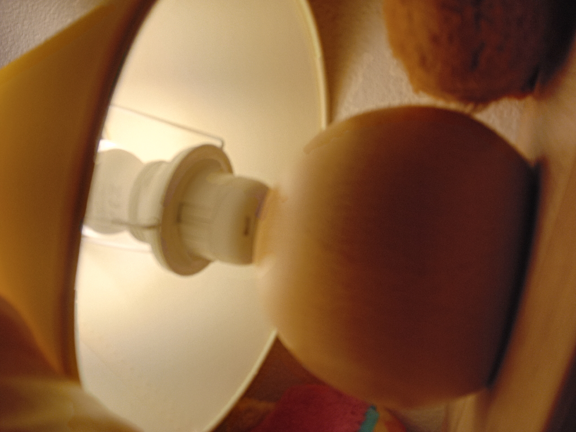 A close-up, low-angle shot of a lamp's base and shade is featured, with the base being a smooth, rounded, light brown object. Above it, the lampshade is a broad, light yellow disc, and a white light bulb is visible beneath it. To the right of the lamp's base is a rough, textured, orange-brown object. The background consists of a blurred, off-white surface. The image does not contain any discernible features to indicate its location in Zuberec, Slovakia.