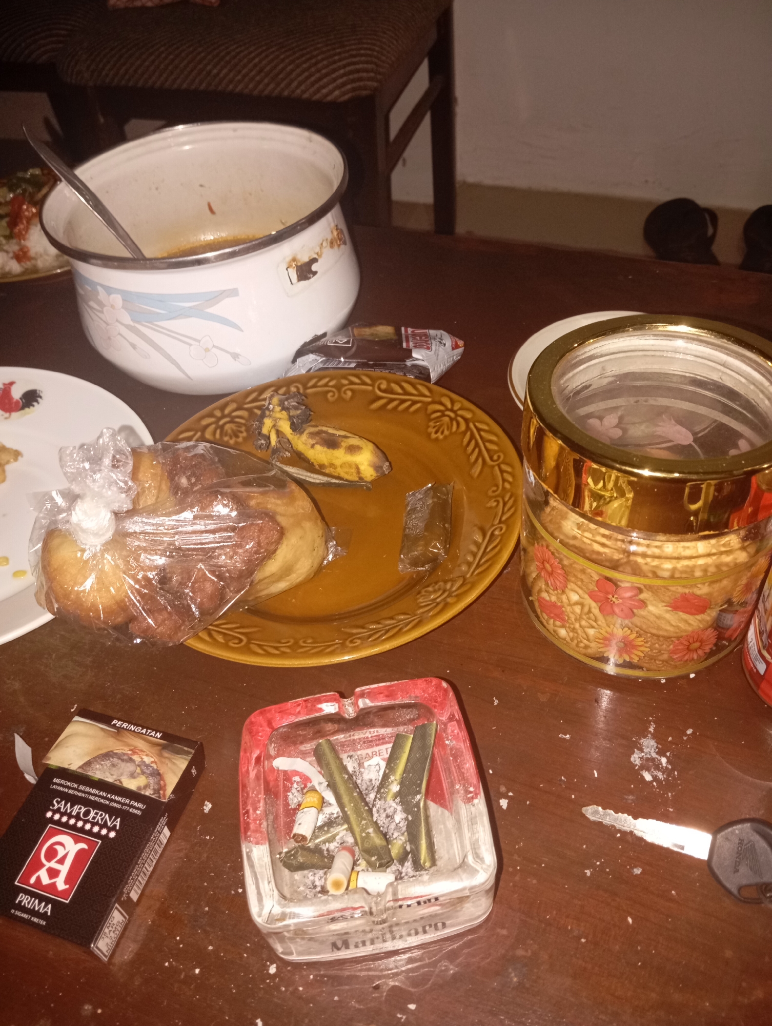 The image depicts a cluttered wooden table in what appears to be a casual indoor setting in Kedungbenda, Indonesia. The table is covered with remnants of food and smoking materials. In the center of the frame, a large white pot with a floral design contains a small amount of leftover food and a serving spoon. To the right of the pot is a brown plate with a half-eaten banana and a wrapped pastry. A decorative glass jar filled with cookies sits to the right of the plate. In the foreground, a pack of Sampoerna cigarettes and a glass ashtray filled with cigarette butts and ash are prominently displayed. A car key lies near the ashtray. In the background, a portion of a woven chair and a wall are visible. The overall impression is one of a post-meal or late-night scene, with evidence of both consumption and smoking.