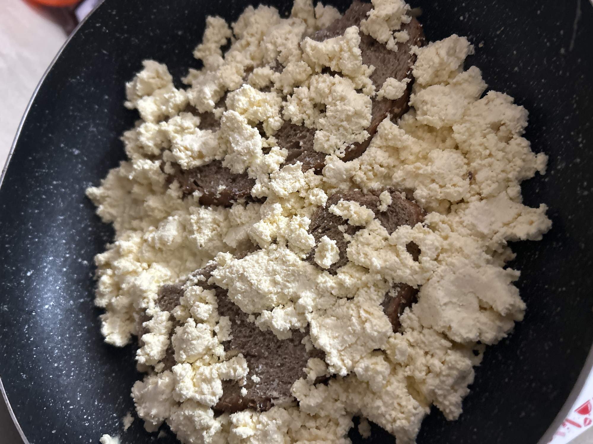The image provides a close-up, top-down view of food being prepared in a dark cooking pan, likely a frying pan. The main subjects are two distinct food items. A base layer of dark brown, possibly reddish-brown, substance is visible underneath. This base is largely covered by a substantial amount of white, crumbly, and lumpy food. The white substance has an irregular, granular texture, strongly suggesting it is a type of fresh cheese, such as cottage cheese, crumbled feta, or a Bulgarian curd cheese like sirene or izvara. The darker ingredient beneath could be minced meat, sausage, or a dark-colored dough.
The scene depicts an activity of food preparation or cooking. The white cheese appears to have been recently added on top of the darker ingredient. The pan is dark with light speckles, typical of non-stick cookware. No people are visible, and therefore no emotions can be discerned. The lighting suggests an indoor setting. There are no specific visual cues within the image itself to indicate the provided location of Zlatograd, Bulgaria, nor is there any visible text. The time of day and weather cannot be determined from the image.