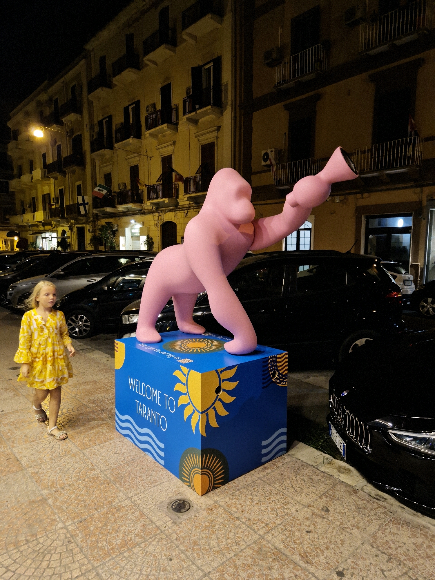 The image depicts a street scene at night in Taranto, Italy, with a prominent pink sculpture of a creature, possibly an ape, standing on a blue cubical base adorned with sun and wave graphics and the text "WELCOME TO TARANTO". A young girl in a yellow dress with bell sleeves and sandals walks on the patterned pavement to the left of the sculpture. Behind the girl and sculpture, several cars are parked along the street. The background features a multi-story building with balconies and windows, illuminated by streetlights, suggesting an urban environment.