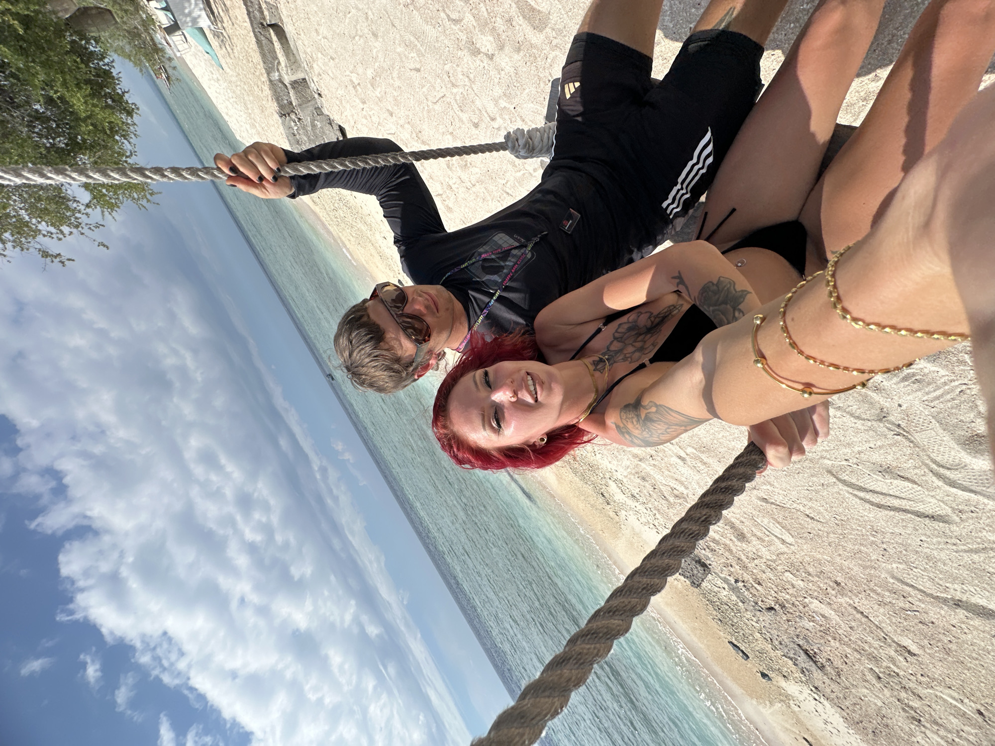 Observed in the city of Unknown, Indonesia, a daytime scene depicts two adults positioned horizontally against a backdrop of a beach, ocean, and sky. One individual, sporting sunglasses and a dark, long-sleeved shirt with black shorts, is lying on the beach. Adjacent to this person lies another individual with red hair, tattoos, and gold bracelets on their arm. Both figures appear to be holding onto a thick rope. The environment includes the light sand of a beach and the blue hues of the ocean, which meet a sky dotted with clouds.