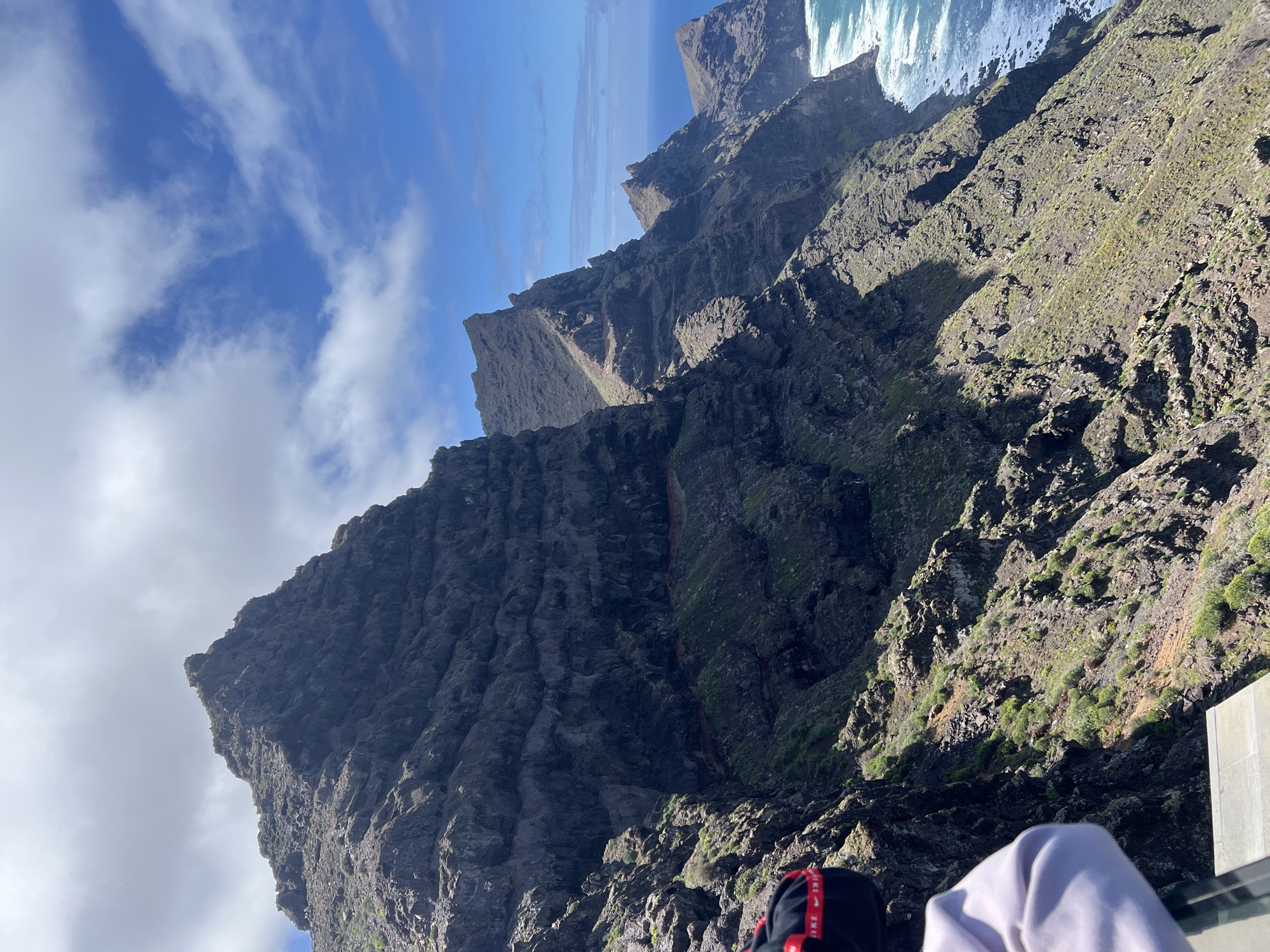 The image depicts the rugged coastline of Tirma, Spain. Jagged cliffs dominate the scene, extending towards the blue sky with scattered clouds. The rocky terrain is textured and covered with sparse vegetation. The ocean is visible in the distance, with waves crashing against the base of the cliffs. Parts of a person is visible, the person wearing a white long-sleeved garment, and a black bag with red stripes. The image is taken from a high vantage point.