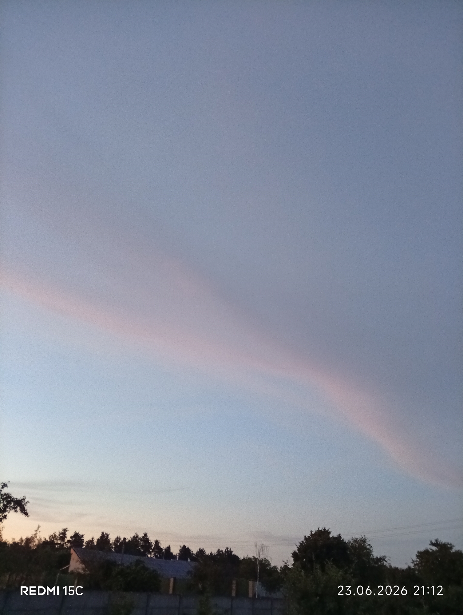 The image captures a serene evening sky over a rural landscape in Mykolaivka, Ukraine. The primary subjects are the sky and the horizon. A distinct, thin cloud formation stretches diagonally across the upper half of the frame, illuminated with soft hues of pink and pale orange, suggesting the presence of the setting sun. The lower portion of the image reveals a silhouette of trees, interspersed with hints of buildings, possibly houses and a fence.
The time of day is clearly indicated as evening, specifically 21:12 on June 23, 2026, as text overlays on the image denote. The sky is a gradient of soft blues and grays, transitioning into the warmer tones near the horizon, indicative of twilight. The weather appears calm, with minimal cloud cover except for the prominent pink-tinged band. The location is identifiable as Mykolaivka, Ukraine, through the provided context.
Visible text includes "REDMI 15C" in the bottom left corner, likely the model of the phone used to capture the image, and "23.06.2026 21:12" in the bottom right corner, indicating the date and time. The overall mood conveyed is peaceful and tranquil, a common feeling associated with the end of a day in a quiet, natural setting. No people or specific objects are clearly discernible in the foreground, allowing the focus to remain on the atmospheric display and the quiet landscape.