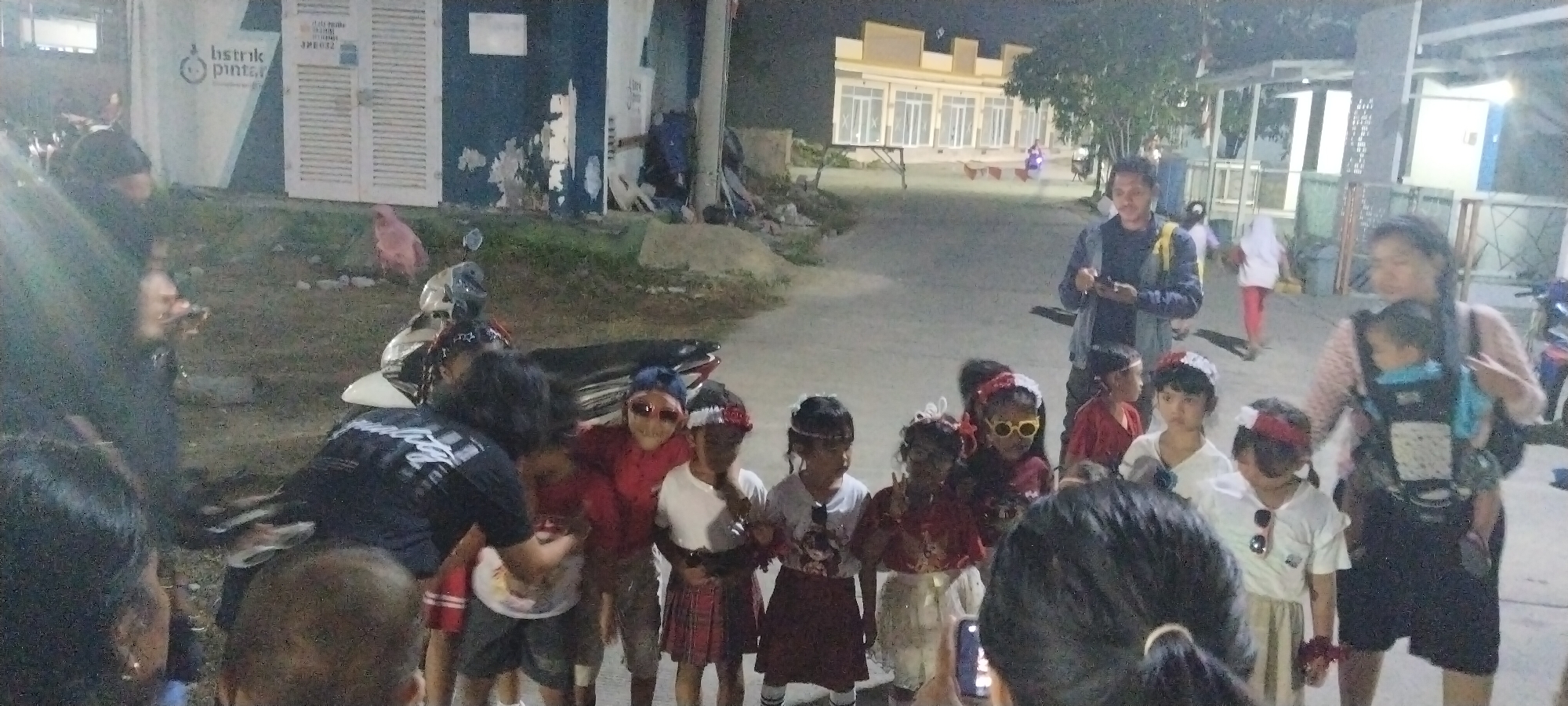 The image depicts an outdoor scene at night, likely in the city of Taban, Indonesia, where a group of people, predominantly children, are gathered. The children appear to be dressed up, some wearing headbands, sunglasses, and festive clothing, suggesting a celebration or performance.
In the foreground, several children are clustered together. One child in the center is wearing a red shirt and large, reflective sunglasses. To their left, a child in a white shirt and a red and white headband stands looking forward. Another child, also with a headband, is visible to the right.
Behind the children, an adult male is standing and holding what appears to be a mobile phone. He is wearing a dark jacket and a yellow backpack. To the right of the male, an adult female is seen carrying a baby in a carrier on her chest. She is wearing a patterned top.
Further in the background, a building with a light-colored facade and windows is visible, along with trees and streetlights that illuminate the scene. There are also several parked motorcycles, one of which is white and located to the left of the children. In the far background, other individuals can be seen walking.
The environment appears to be a paved area, possibly a street or courtyard, with some grassy areas and debris on the ground. The overall impression is of a community gathering, likely an event for children, taking place after dark.