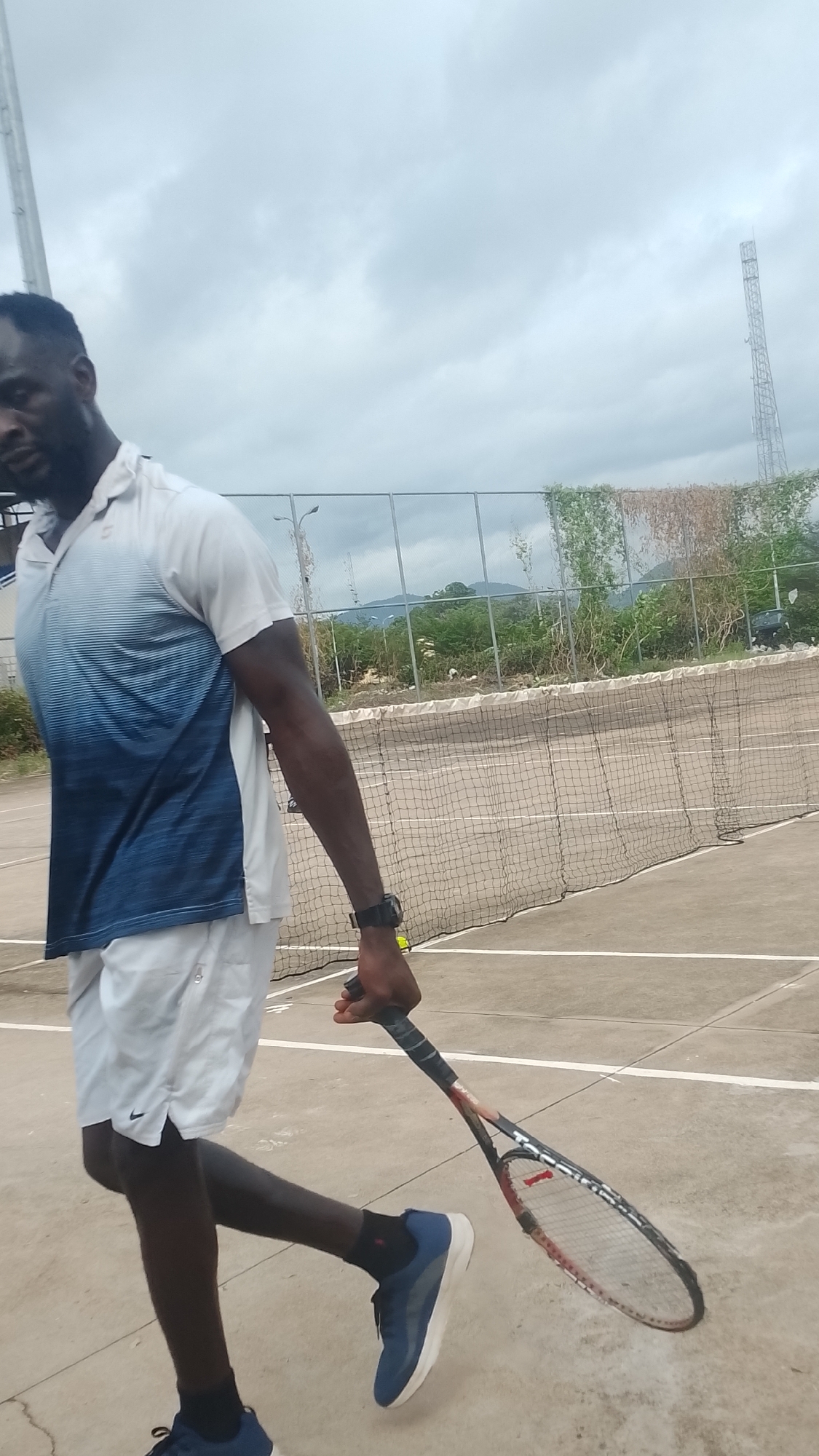 An outdoor scene in Lokoja, Nigeria, features an adult male walking on a hard-surface tennis court under an overcast sky. The man, with dark skin, is in the left foreground, mid-stride with his left leg bent and right leg extended. He wears a white polo shirt with a blue gradient pattern on the lower half, white athletic shorts (displaying a Nike logo on the left thigh), black socks, and blue and white athletic shoes. A black wristwatch is on his left wrist. He holds a red and black tennis racket in his right hand, with the racket head angled towards the ground. A single yellow tennis ball is visible on the court surface near the net, slightly behind his right hand. In the mid-ground, a black tennis net with a white top band stretches across the court. Behind the net, a metal fence separates the court from a background containing green and dry vegetation, utility poles, and distant mountains. A tall metal communication tower is prominently visible on the far right horizon, extending into the cloudy sky. The tennis court surface is light-colored concrete with white boundary lines.
