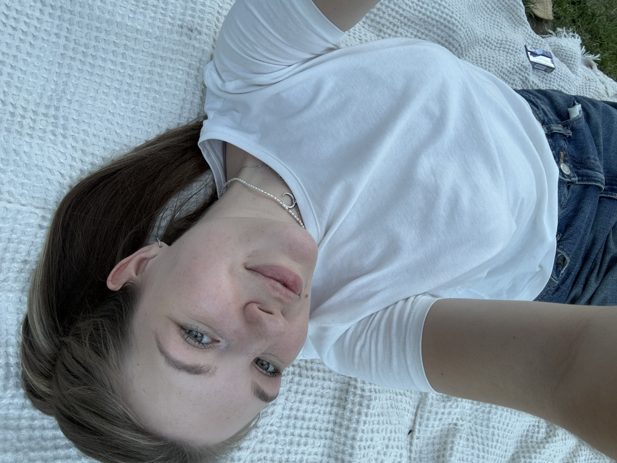 A young woman lies on a white textured blanket outdoors. She wears a white t-shirt, blue jeans, and a silver necklace with a circular pendant. Her hair is pulled back in a ponytail. A pack of cigarettes sits on the blanket near the upper right corner of the image. The location is unspecified, but it is possibly in the Cukarica Urban Municipality, Serbia.
