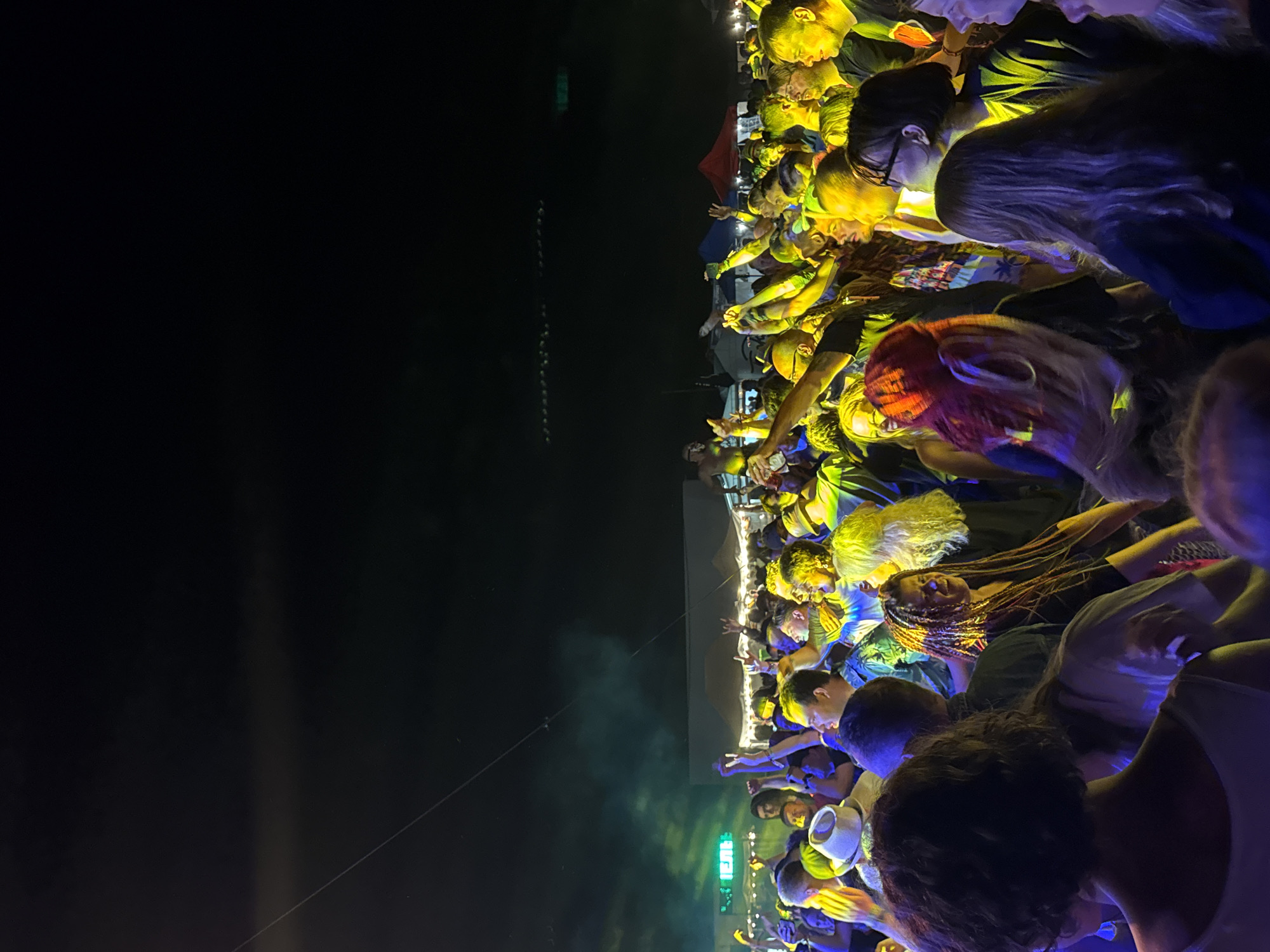 The image depicts a nighttime outdoor event, possibly a music festival or concert, in an urban setting, likely Unknown, Unknown. A large crowd of people are gathered, illuminated by stage lighting that casts a vibrant yellow and green glow. Many individuals are raising their hands in the air, suggesting active participation and enjoyment. The background shows a dark sky with some distant lights and what appear to be tents or temporary structures. The foreground is filled with the faces and upper bodies of concertgoers, some of whom are wearing hats and have distinctive hairstyles. The atmosphere is energetic and festive.