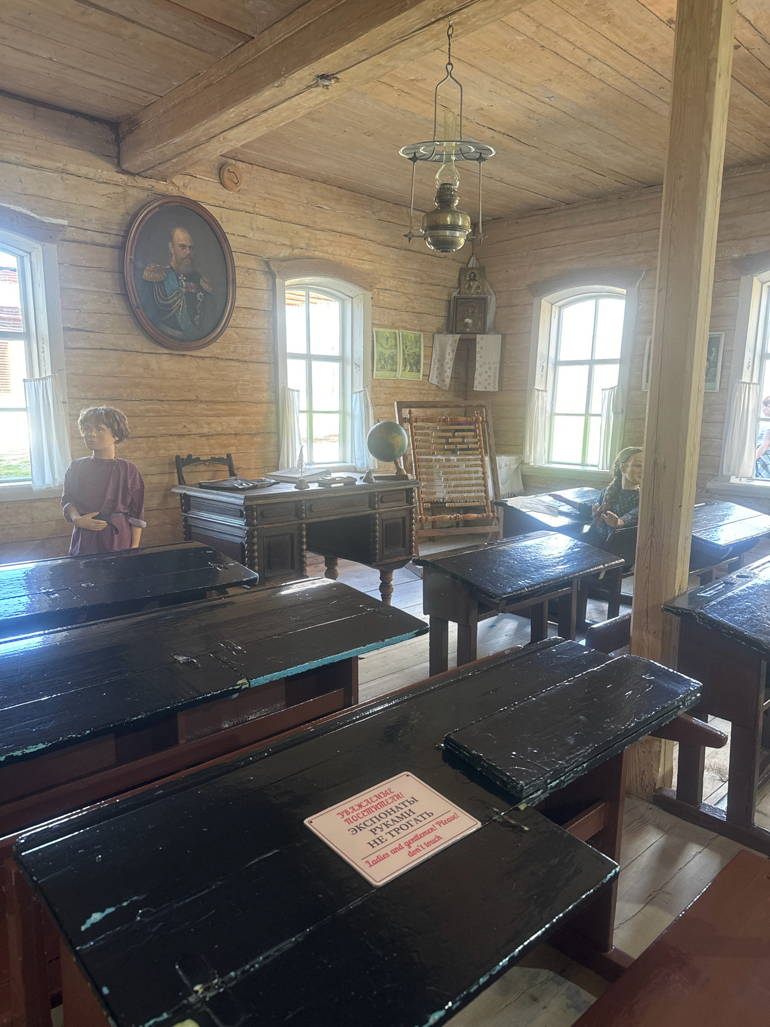 The image depicts a historical classroom setting, likely at the "Taltsy" Museum in Russia, designed to resemble a 19th-century school. The room is constructed with rustic wooden walls and a wooden plank ceiling, with exposed wooden beams. Natural light enters through large, multi-paned windows draped with simple white curtains.
In the foreground, several rows of old-fashioned wooden school desks are visible. These desks are painted black, with some showing signs of wear. A small, pink sign with text in Russian and English reading "Ladies and gentlemen! Please! don't touch" is affixed to one of the desks.
On the left side of the room, a mannequin dressed in a traditional dark purple garment stands near a window. To the right, another mannequin, this one appearing to be a young girl with braided hair, is seated at a desk.
Further back, a large, ornate wooden teacher's desk is positioned with a chair behind it. On the desk, a globe, an abacus, and writing implements are arranged. A framed portrait of a distinguished-looking man in military attire hangs on the wall above the teacher's desk. A hanging oil lamp is suspended from the ceiling. Several framed pictures and religious icons are also visible on the walls.
The overall impression is of a preserved historical interior, offering a glimpse into the educational environment of a past era.