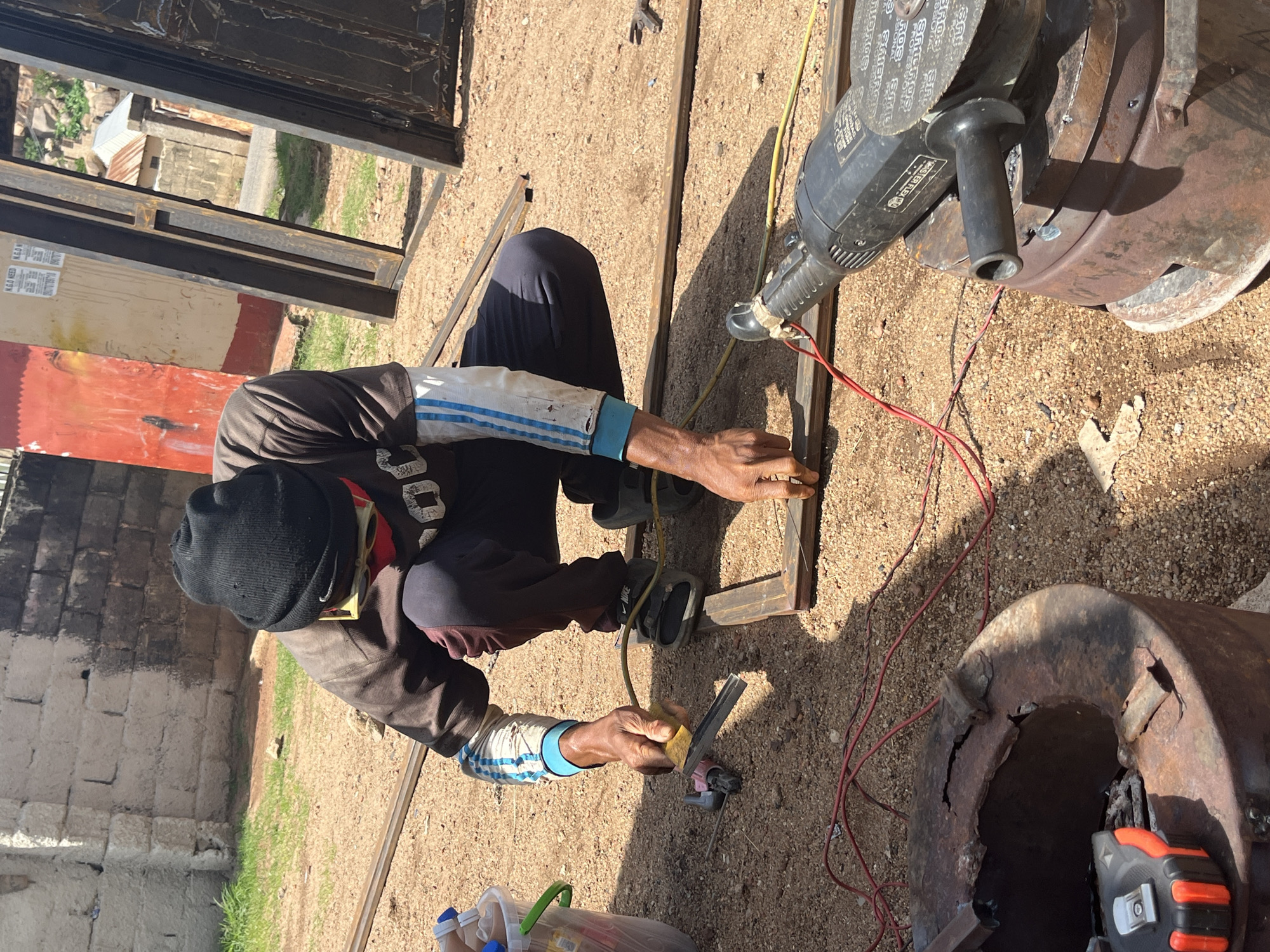A lone male welder, wearing a black beanie, a brown long-sleeved shirt with blue and white stripes on the arms, dark pants, and red safety glasses, is crouched on the ground, working on metal framing. He holds a yellow sanding pad against a piece of metal. A red and black measuring tape, a small grinder, and red electrical wires are visible on the ground near him. In the background, there are large metal doors leaning against a wall, some concrete structures, and vegetation. The scene appears to be an outdoor workshop or construction site. The lighting suggests it is daytime, with bright sunlight casting shadows. The ground is covered with sand and small pebbles. The overall environment suggests a somewhat dusty and utilitarian setting. The image was taken in Jos, Nigeria.