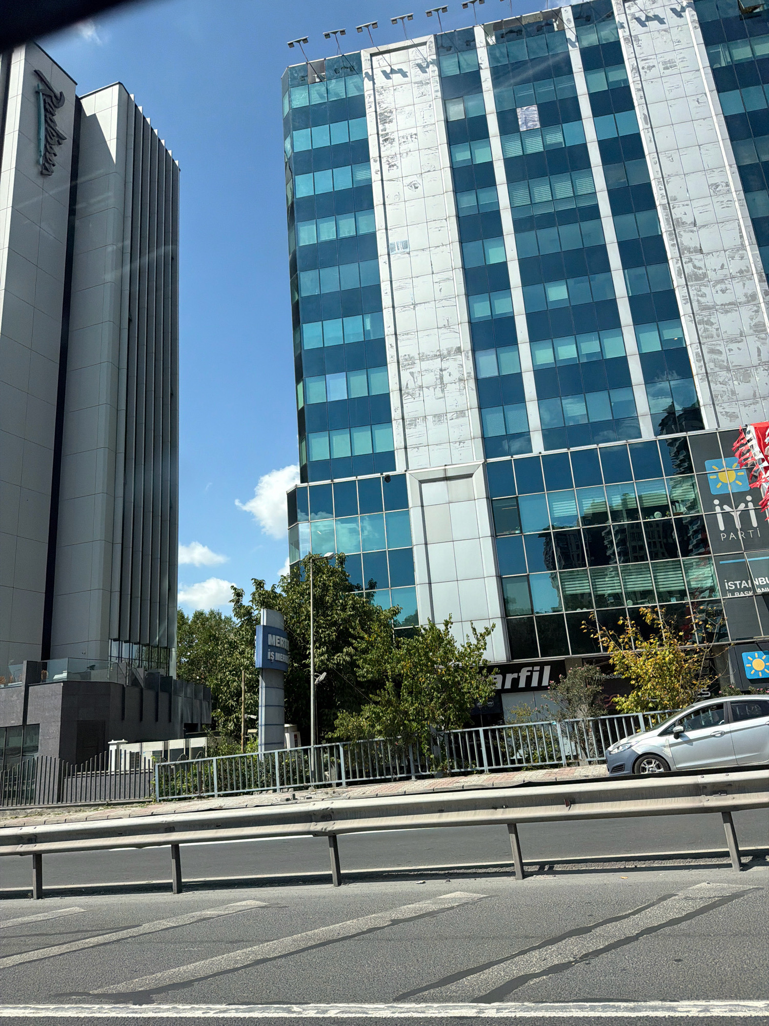 The image shows two modern office buildings in Zeytinburnu, Turkey, on a sunny day with a clear blue sky and scattered clouds. The building on the left is a tall, rectangular structure with a light gray facade featuring vertical ribbed detailing. A prominent sign with the "Radisson" logo is visible on its upper left side. To the right of this building is a taller, glass-fronted skyscraper with a blue tint to its windows. This building has white vertical beams separating the windows, and some sections of its facade appear weathered. Below the glass facade, several signs are visible, including "iYi PARTI" and "İSTANBUL İL BAŞKANLIĞI". In the foreground, a multi-lane road with a metal safety barrier runs horizontally across the image, with a silver car driving on the right lane. Trees and foliage are visible between the buildings and behind the road barrier, some displaying autumn colors. The overall scene depicts an urban environment with commercial architecture.