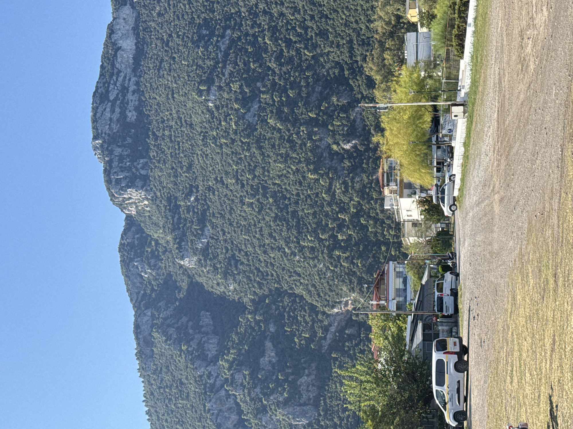 The image shows a rocky mountain densely covered with trees, dominating the background. In the foreground, a parking area is visible with a gravelly surface and patches of dry grass. Several vehicles are parked in the area, including a white van with yellow stripes, another white van, a silver car, and a motorcycle. Buildings, possibly residences or businesses, are situated at the base of the mountain, some with red-tiled roofs. Lush green trees and lighter green, leafy trees are interspersed among the buildings and along the edge of the parking area. The sky is clear and blue. The scene appears to be a street or access road within the Municipal Unit of Kamena Vourla, Greece, likely a commercial or residential area at the foot of a mountain.