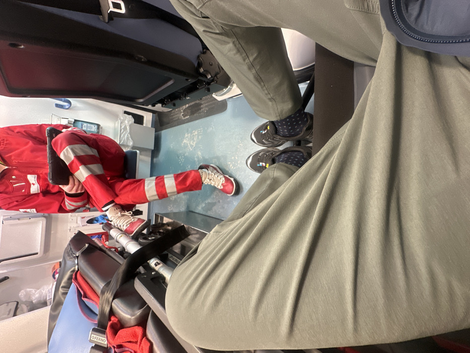 Inside an ambulance, a person in a red uniform with white stripes sits, holding a black tablet. Beside them, someone in olive green pants and dark sneakers with patterned socks sits casually. The interior reveals standard ambulance equipment and seating. The perspective is from a high angle, focusing on the lower body of both individuals.