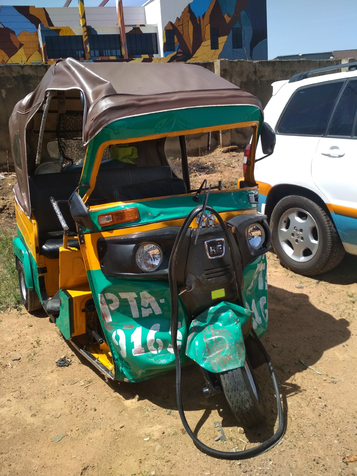 The image shows a yellow and green Bajaj Qute parked on a dirt surface. The vehicle has a brown roof and "PTA 916" painted in white on its front. A white SUV is parked next to the Qute. The background features a building with a colorful mural. The location appears to be a city in Unknown, Unknown.