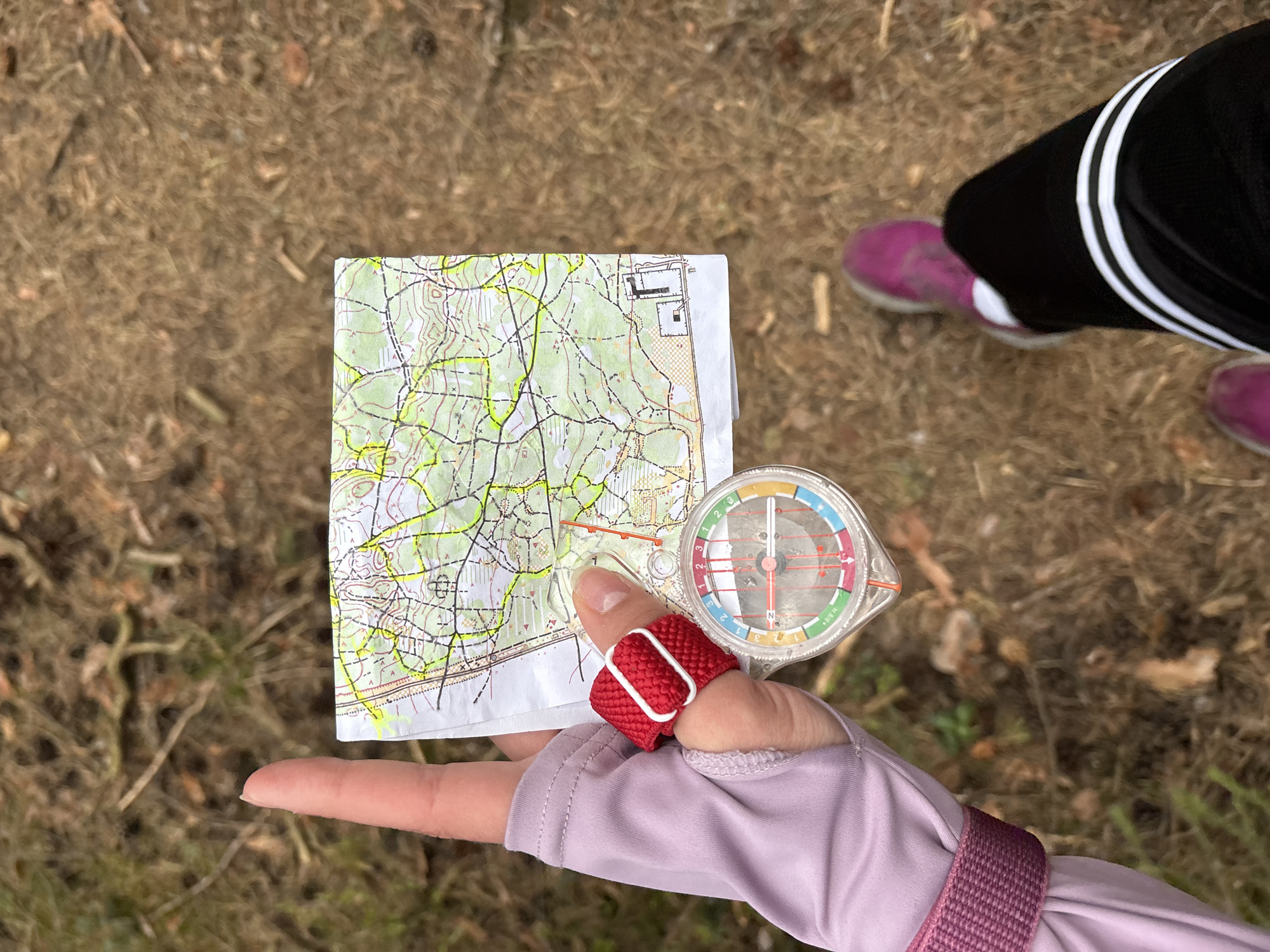 An individual is holding a topographical map and a compass in their left hand. The hand is wearing a purple long-sleeved shirt and has a red, elastic thumb brace with a white plastic buckle. The map is folded and shows terrain features and possibly roads or trails, marked with yellow highlighter. The compass is clear plastic with a colorful dial. The background shows dry grass and leaves, with the lower legs of another person wearing black track pants with white stripes and purple sneakers visible in the upper right corner. The location is not discernible from the image, so it cannot be determined whether this image was taken in Perm, Russia.