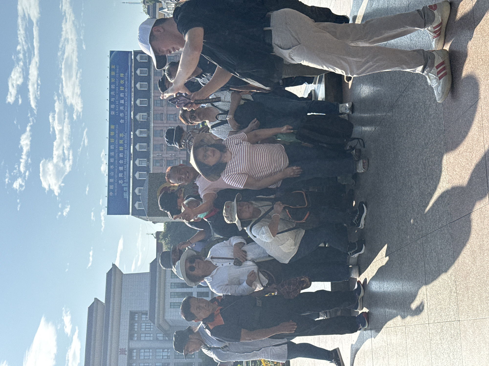 A group of approximately twelve people, predominantly of Asian descent, is gathered on a paved plaza in what appears to be Lao Cai Ward, Vietnam. They are standing and looking up at a large blue billboard displaying text in both Chinese and Vietnamese languages. A man in a baseball cap and athletic shoes seems to be taking a picture of the billboard with his smartphone. Buildings are visible in the background, and the sky is clear with scattered clouds.
