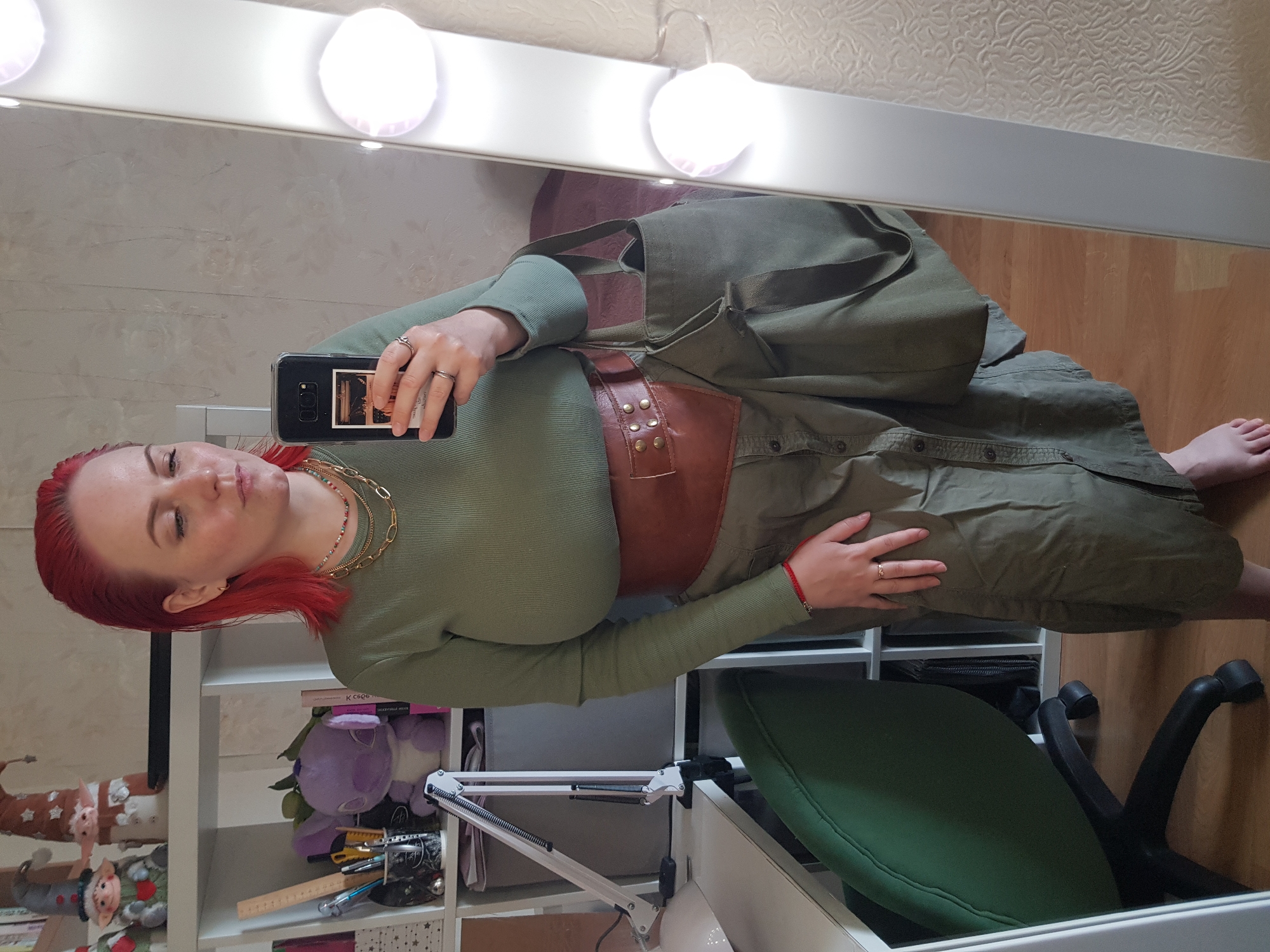 The image depicts a woman with red hair, wearing a long-sleeved, light green shirt and an olive green button-down dress cinched at the waist by a brown leather belt. She is taking a selfie using a black smartphone. The setting appears to be indoors, likely a bedroom or dressing room. A shelf unit filled with various items, including stuffed animals, is visible in the background. A green office chair is partially visible in the lower right corner of the image. The lighting is provided by a row of bright vanity lights above a mirror, reflecting the woman and the scene. The location is indicated as Unknown, Unknown.