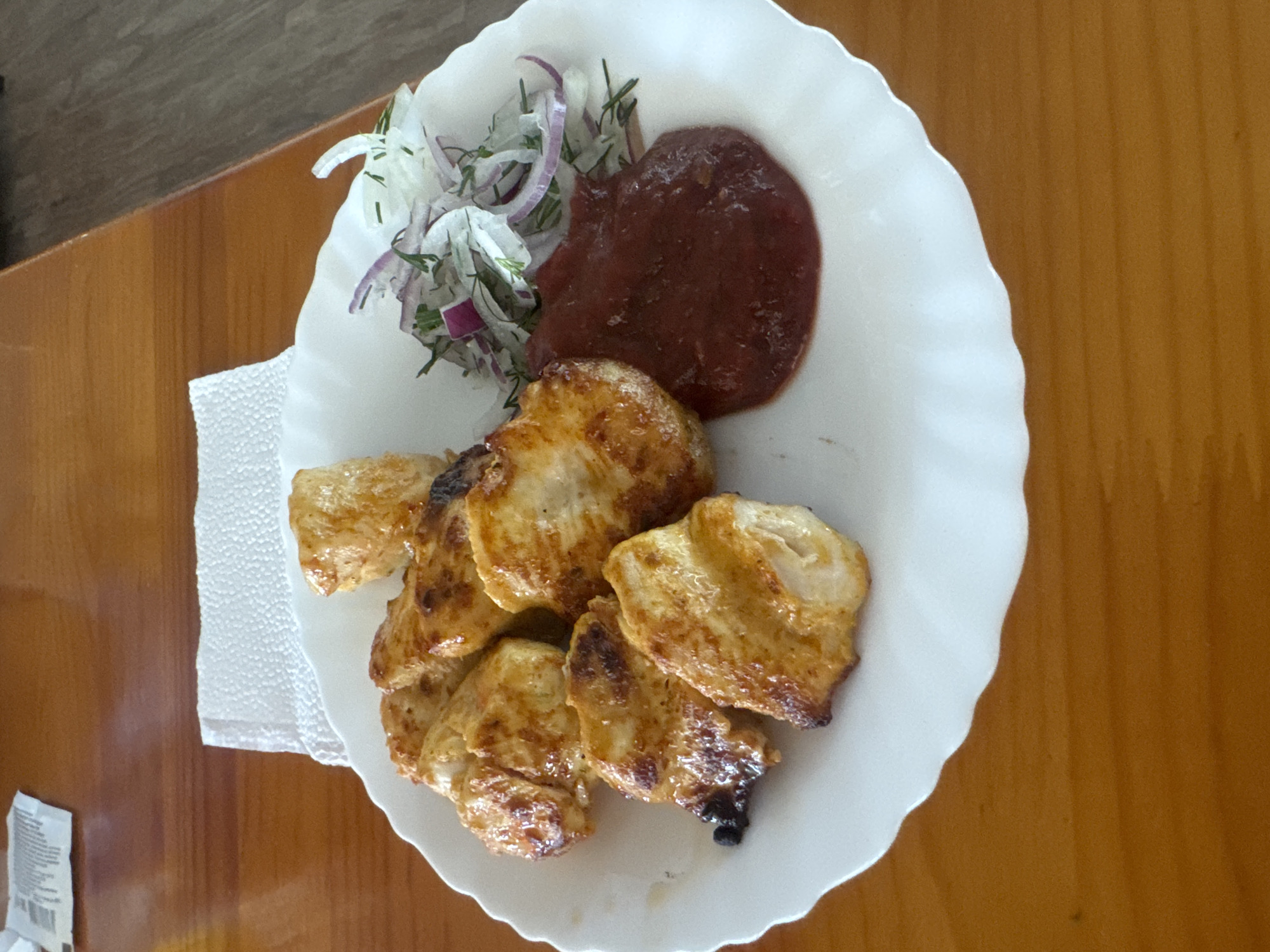 A white plate of food sits on a wooden table. The plate contains several pieces of golden-brown grilled chicken, a dollop of dark red sauce, and a small salad of thinly sliced red onion and green herbs. A white napkin is placed behind the plate. A small white packet is visible in the upper left corner of the image. The scene appears to be indoors, with natural light illuminating the food.