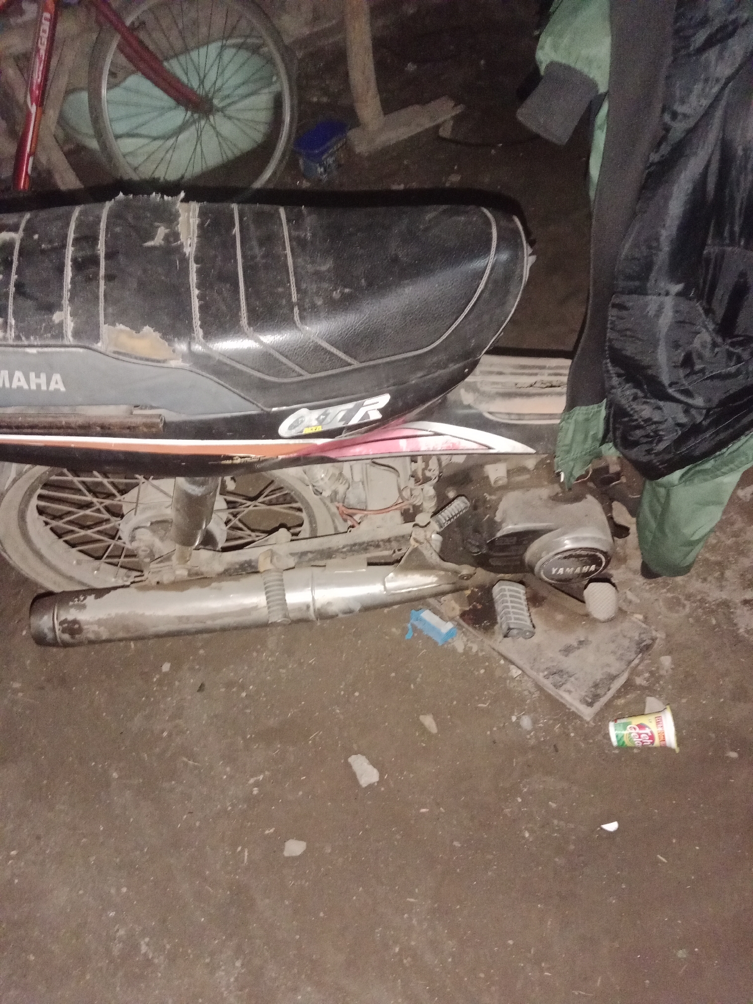 A Yamaha motorcycle seat is visible in the foreground of the image, with the word "YAMAHA" clearly visible in white lettering on the side. The seat appears to be made of black leather and shows signs of wear and tear. The motorcycle's engine is partially visible, also bearing the "YAMAHA" logo. A silver exhaust pipe runs along the bottom of the motorcycle. In the background, a bicycle wheel with red spokes can be seen. The ground appears to be dirt or concrete, with some debris scattered around. The scene is lit with artificial light, suggesting it may be nighttime or an indoor location. The overall impression is one of a possibly abandoned or disused motorcycle and bicycle, likely in an outdoor or semi-outdoor setting in Ringinrejo, Indonesia.