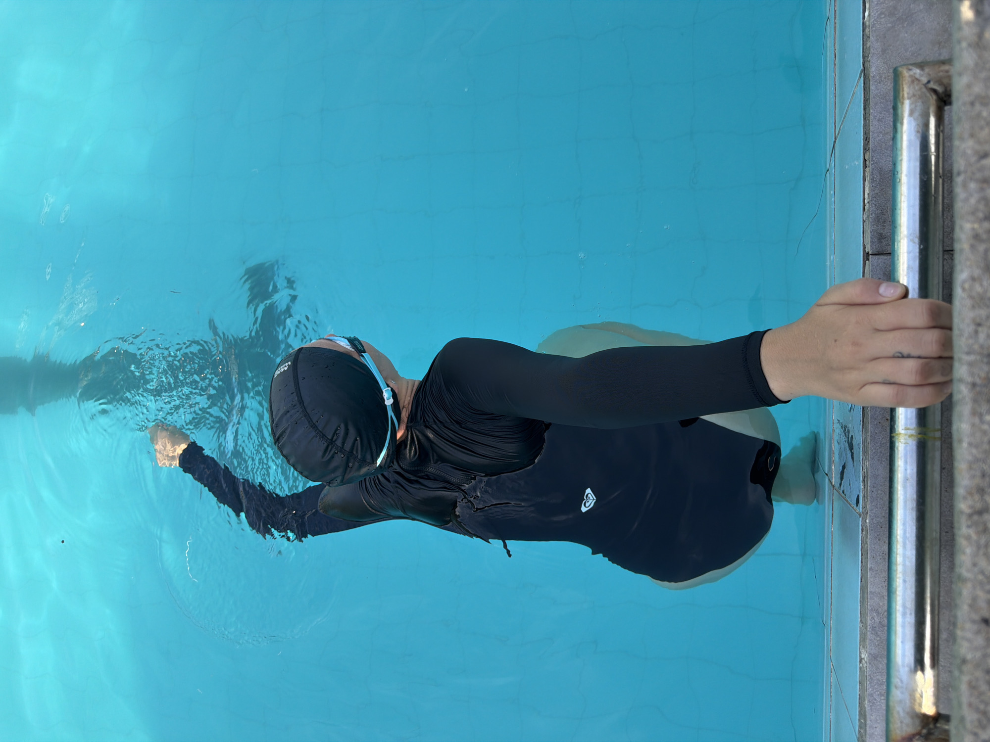 A person wearing a black long-sleeve shirt, black swim cap, and goggles is swimming in a blue-tiled swimming pool. Their left hand is outstretched, while their right hand is holding onto the metal pool ladder. The scene is located in Nha Trang Ward, Vietnam.