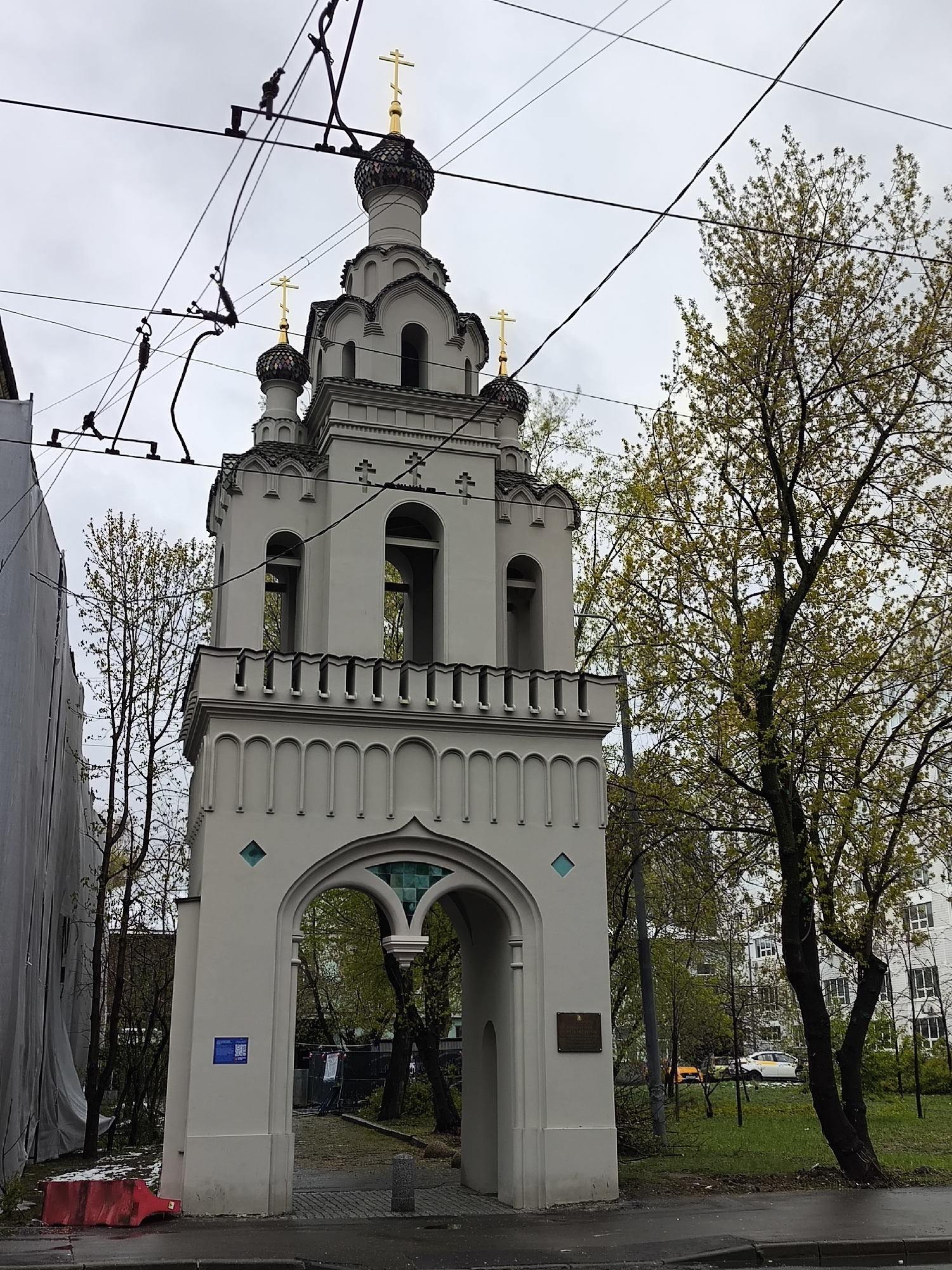 An ornate, multi-tiered bell tower, likely part of an Orthodox church, stands prominently in an urban setting under an overcast sky. The tower is constructed from a light-colored material, possibly off-white or light grey, and features intricate architectural details including arched openings for bells, decorative moldings, and a crenellated upper balcony. It is topped with multiple onion domes, some with a scaled or patterned surface, each crowned with a golden Orthodox cross.
The bell tower's base forms a large arched gateway, through which a green area with trees and a distant building can be seen. Two plaques are affixed to the tower's lower section: a blue sign with a QR code and Cyrillic text on the left pillar, and a darker metal plaque on the right.
Overhead, a network of black trolleybus wires crisscrosses the muted grey sky. To the left of the bell tower, a building is partially covered by white construction sheeting, with a tree exhibiting sparse spring foliage nearby. On the right, a large tree with an abundance of vibrant, light green leaves, indicative of springtime, dominates the midground, partially obscuring a multi-story residential building and a white car with a yellow stripe (possibly a taxi) in the distance. A red plastic barrier rests on the pavement in the immediate left foreground. The scene depicts an urban landscape during daytime, with no human activity visible, under cloudy weather conditions. The architectural style suggests the location is Moscow, Russia.