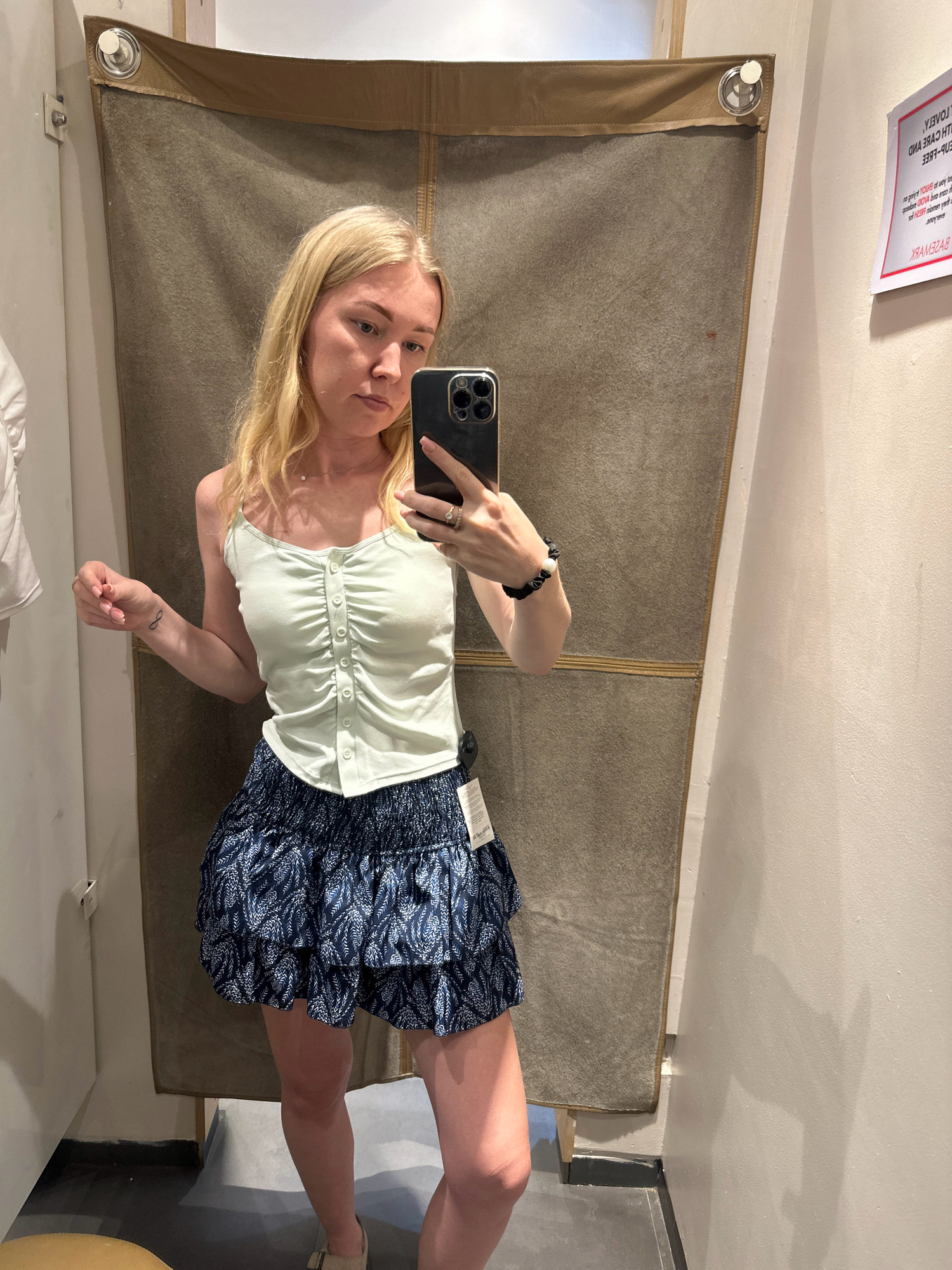 A young woman with blonde hair is taking a mirror selfie in a fitting room. She is wearing a light green ruched camisole with buttons and a blue and white patterned ruffled skirt. She has a black scrunchie on her wrist and is wearing light-colored flat shoes. A white tag is visible hanging from the skirt. The fitting room has a textured brown curtain and a light-colored wall. A sign is posted on the wall to the right of the woman. The scene appears to be in a retail store, possibly in Kathmandu Metropolitan City, Nepal, given the context of the request.