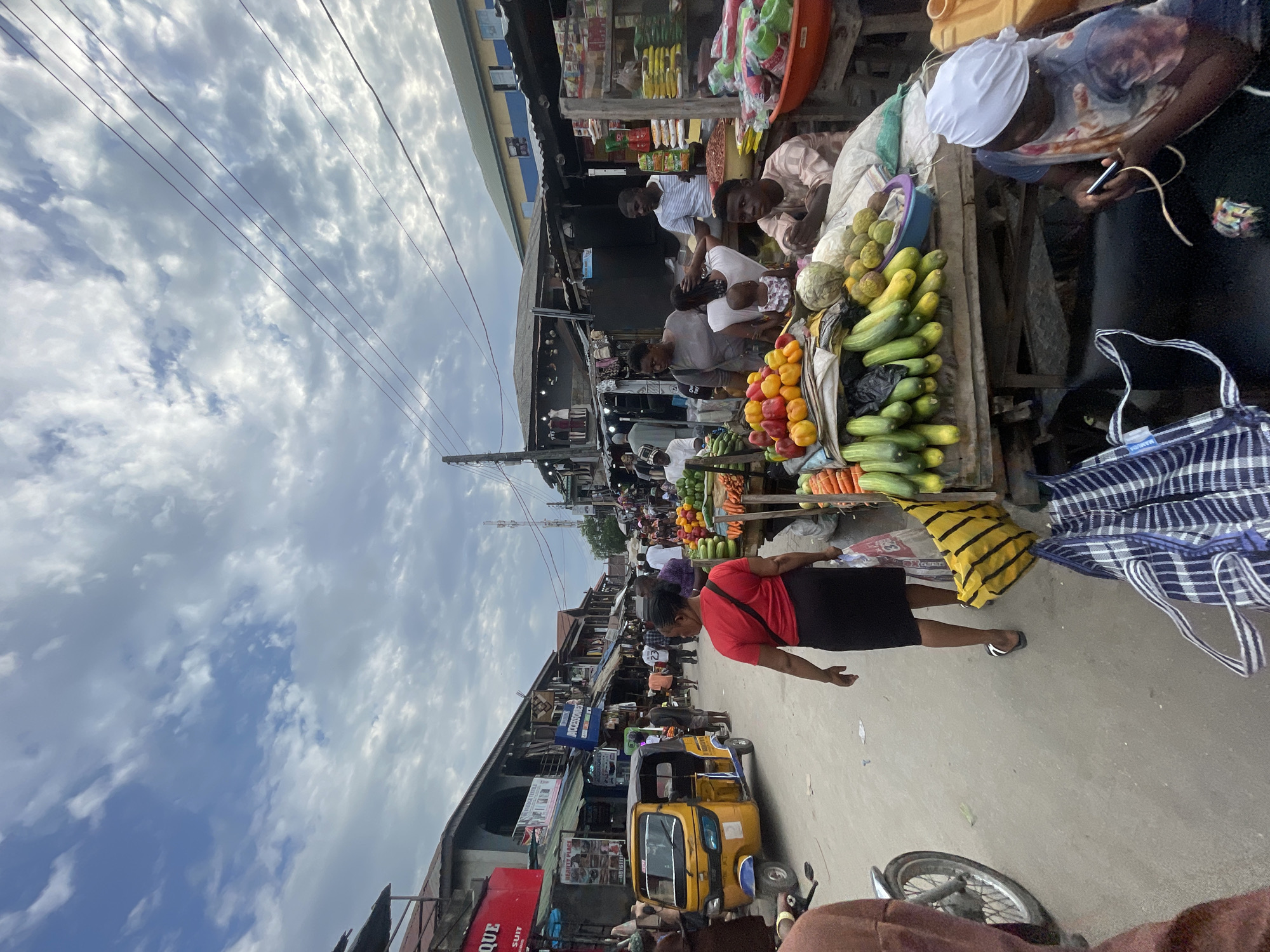 The image depicts a bustling open-air market in what is likely Ado, Nigeria, based on visual cues. Several vendors display goods, including fruits and vegetables like cucumbers, oranges, and carrots on tables. People are shopping and interacting with the vendors. A woman in a red shirt and black skirt is walking in the foreground, while a yellow "keke" or auto-rickshaw is parked nearby. The market stretches into the distance, with numerous people and stalls visible. Power lines crisscross the sky above. Buildings line the street, with shops advertising various goods and services.