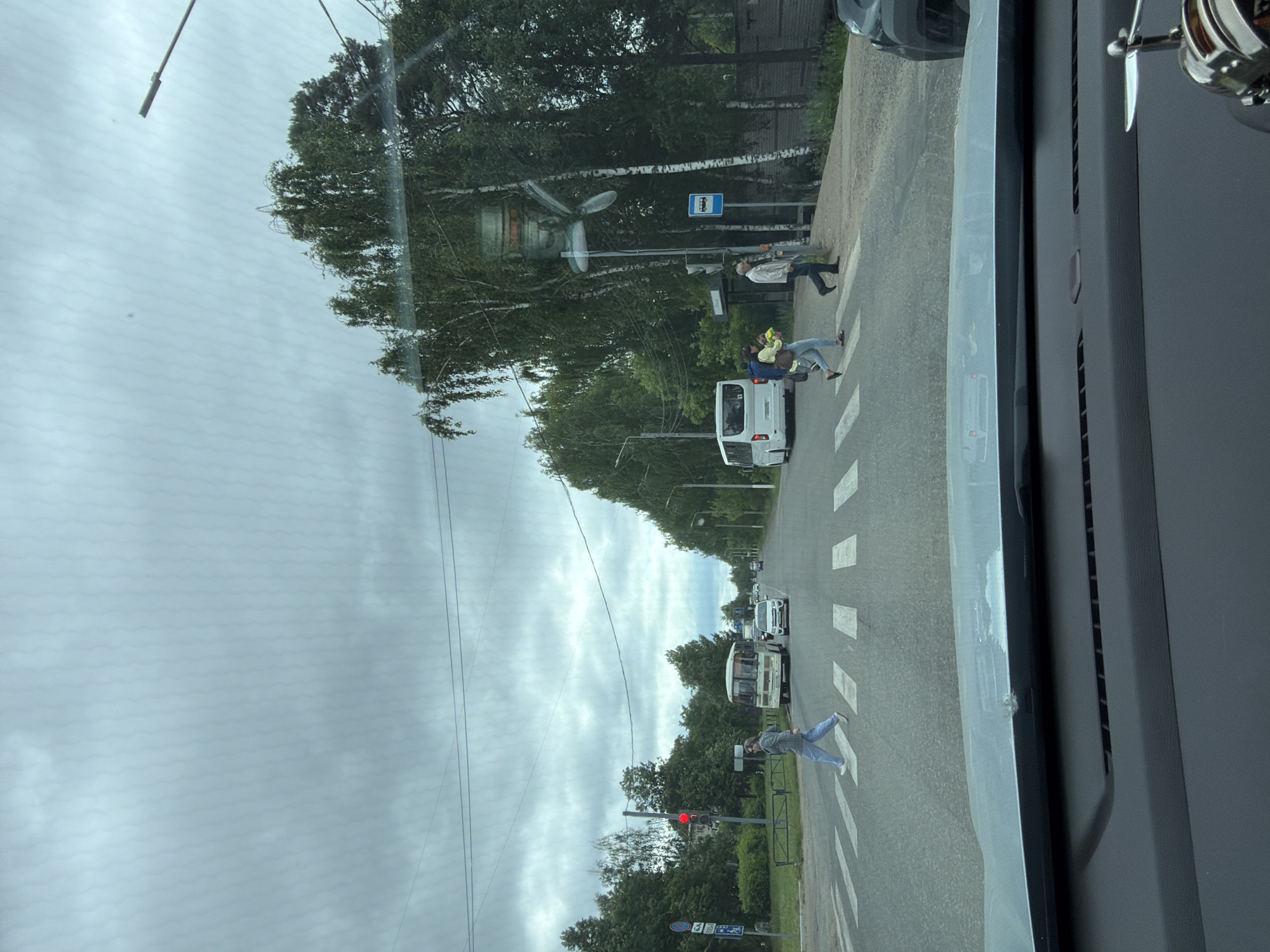 This is a ground-level, eye-level, medium shot of a cloudy day at a crosswalk in Chaikovsky, Russia. The scene is captured from inside a vehicle, looking out through the windshield, which shows reflections of the sky and trees. The crosswalk is marked with white stripes on a gray asphalt road.
On the left side of the crosswalk, a traffic light shows red. A woman in blue jeans and a gray jacket is walking across the crosswalk from left to right. A white bus is stopped at the crosswalk behind her, and a white car is behind the bus.
Further down the road, on the right side, two people are crossing the crosswalk. A woman in a yellow top and blue jeans is holding a bag, and a man in a light-colored jacket and dark pants is walking behind her. A white van is visible on the road beyond them, and trees line the right side of the street. A bus stop sign is also visible near the right sidewalk.
In the foreground, the dashboard of the vehicle is visible, with a silver air freshener with propellers on the right side. The overall atmosphere is overcast and quiet, with signs of normal urban activity.