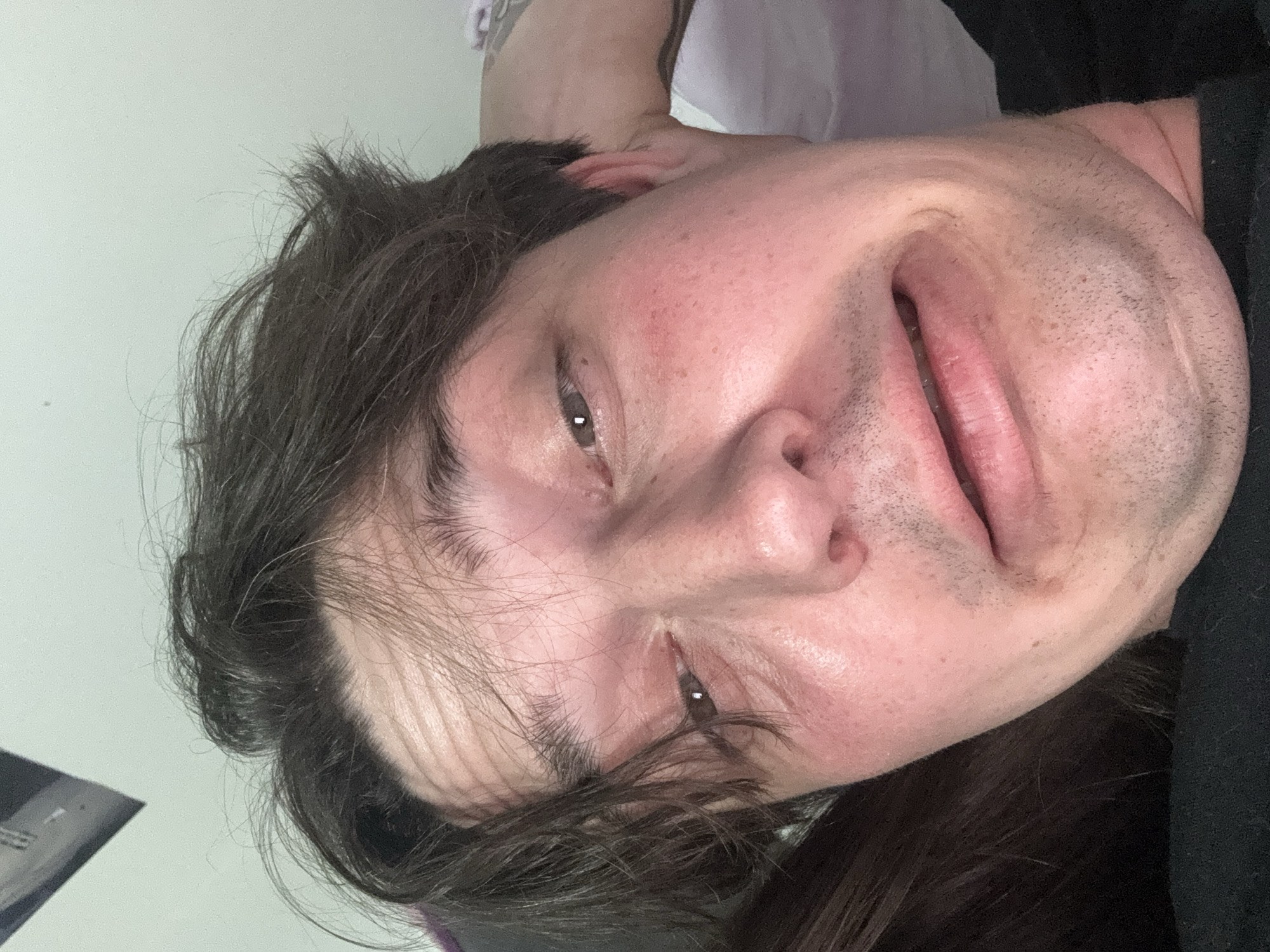 This is a close-up selfie of a person with tousled brown hair and fair skin, looking directly at the camera with a strained or uncomfortable expression. Their eyes are open, and their mouth is contorted into a grimace. The lighting suggests an indoor setting, possibly a bedroom, with a pale green wall in the background. There are some shadows on the forehead, creating horizontal lines. The person is wearing a black garment, and part of another person's dark hair is visible on the left side of the frame. A portion of a tattoo is visible on the person's right arm. The image does not contain any information about the location in Kyiv, Ukraine.