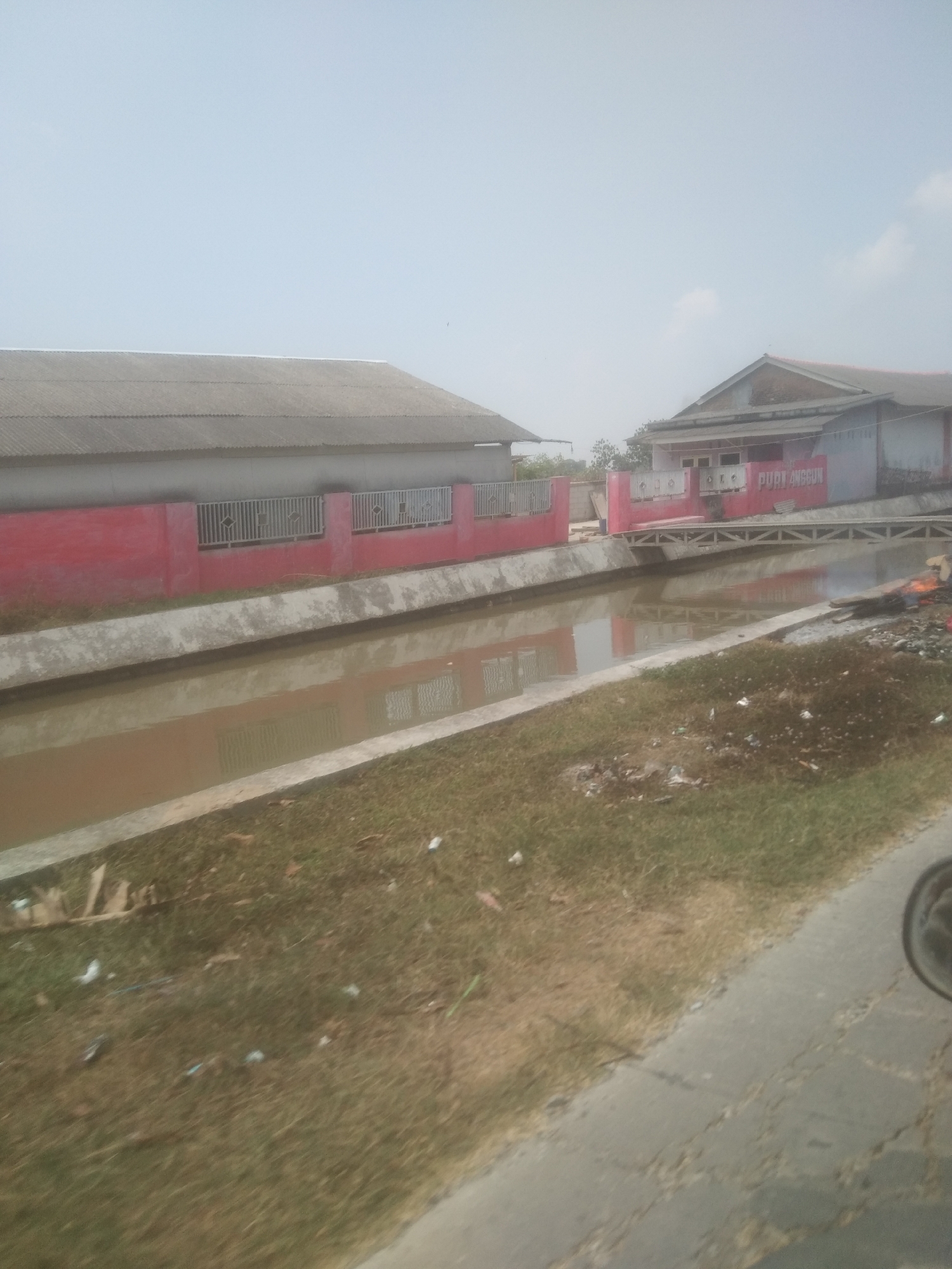 The image captures a canal running through a semi-urban area in Desa Cilandak Lor, Indonesia. On the left side of the canal, a long, single-story building with a gray corrugated roof and a vibrant red lower wall is visible. This building features several rectangular metal-grated windows. Across the canal, a smaller, two-story building with a reddish-brown tiled roof and white walls is situated. This building has the text "PURI ANGGUN" written in red lettering on its facade. A narrow metal footbridge spans the canal, connecting the two sides. The canal itself appears to be filled with murky brown water, reflecting the surrounding structures. The banks of the canal are lined with concrete and overgrown with grass and scattered debris, suggesting a lack of regular maintenance in some areas. The overall scene is illuminated by daylight under a hazy sky, and the perspective suggests the photo was taken from a moving vehicle.