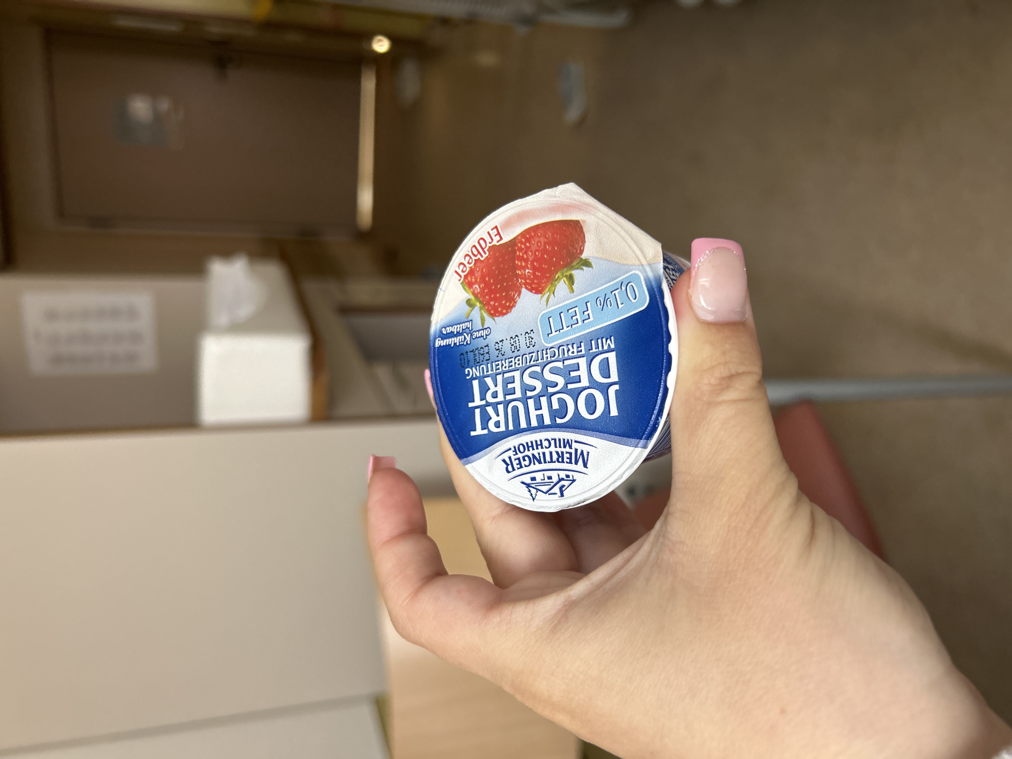 A person is holding a cup of "JOGHURT DESSERT" with strawberries, labeled "Erdbeer" (strawberry) in German. The yogurt is a low-fat variety, indicated by "0,1% FETT". The packaging also states "MIT FRUCHTZUBEREITUNG" (with fruit preparation) and "ohne Kühlung haltbar" (shelf-stable without cooling). The background appears to be an indoor hallway, possibly in a hospital or care facility, with a closed door and a notice on the wall. The lighting suggests an artificial indoor environment. The image is taken from a close-up perspective, focusing on the hand and the yogurt cup, with the background blurred. This setting does not provide any specific indicators to determine a location within the city of Gummersbach, Germany.