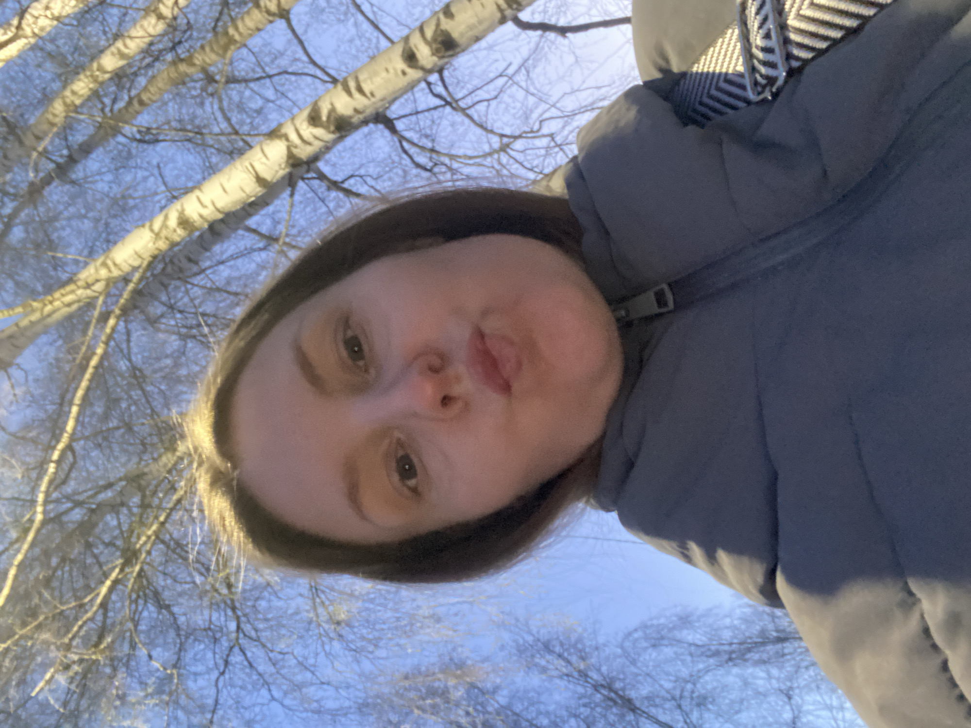 A close-up, upward-angled selfie captures a young woman outdoors against a backdrop of bare tree branches and a light blue sky. The woman, positioned at the bottom of the frame, looks up towards the camera with a subtle puckered lip expression, indicative of a playful pose. Her eyes are open, and she has light-colored hair visible around her face.
She is dressed in a dark gray or blue puffy winter jacket, suggesting cold weather. A striped, chevron-patterned strap from a bag or accessory is visible on her right shoulder area. The background is dominated by a dense network of leafless tree branches, many of which exhibit light, possibly white, bark, characteristic of birch trees found in regions like Noviy, Russia. The sky above is a clear, soft blue, indicating either early morning, late afternoon, or twilight, with diffused natural light illuminating the scene. The overall impression is that of a clear, cold day in late autumn, winter, or early spring. No visible text is present in the media.