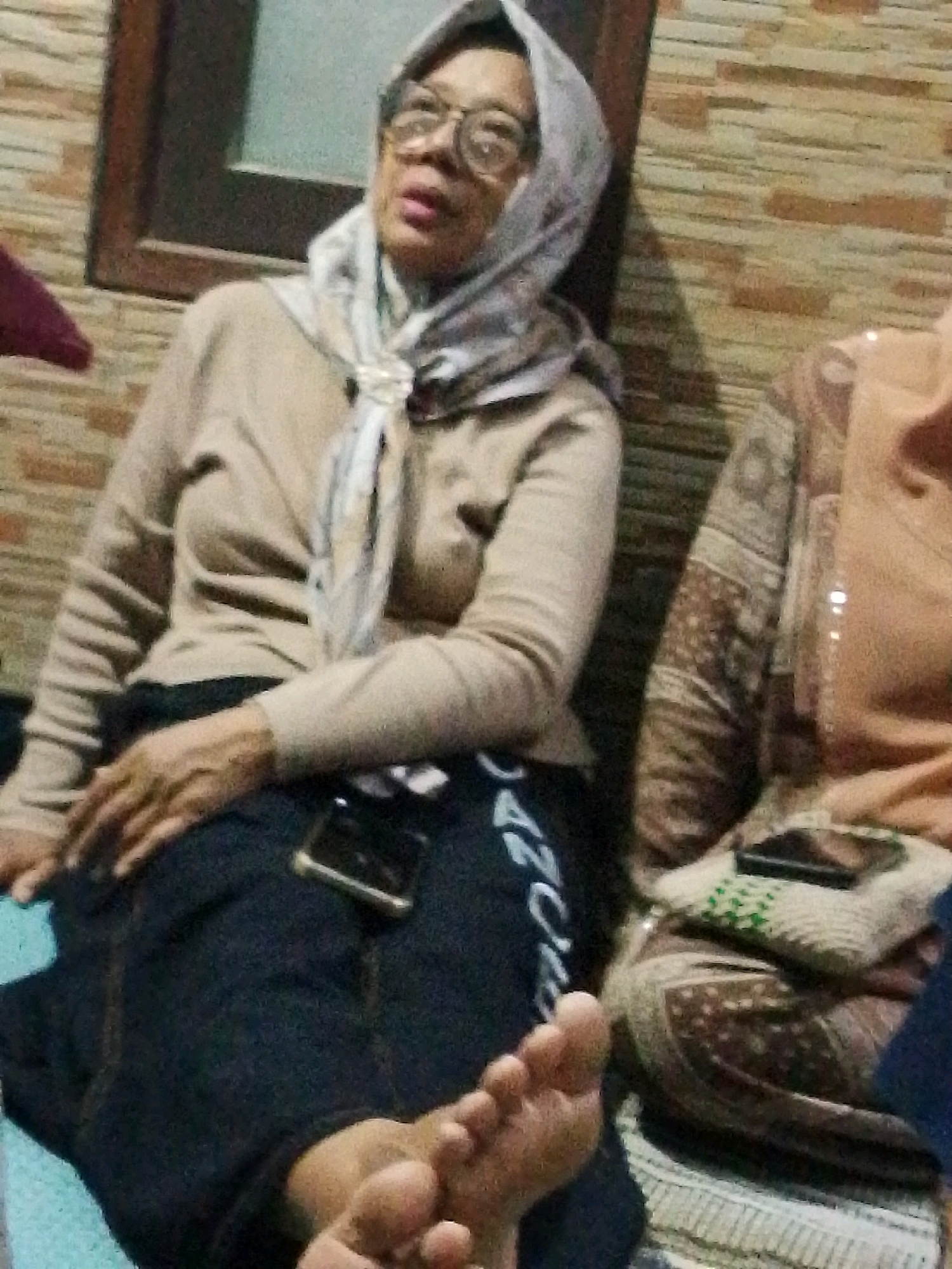In the city of Bulurejo, Indonesia, a medium shot captures two elderly women sitting indoors. The woman on the left, wearing a light brown hijab and glasses, is dressed in a tan sweater and dark pants. Her right hand rests on her lap, and her bare feet are visible in the foreground. She appears to be looking to her left, away from the camera. To her right, another woman, partially visible, is seated and wearing a beige outfit. A patterned cushion lies between them. The background features a brick wall and a dark wooden frame. A smartphone is placed on the woman's lap.