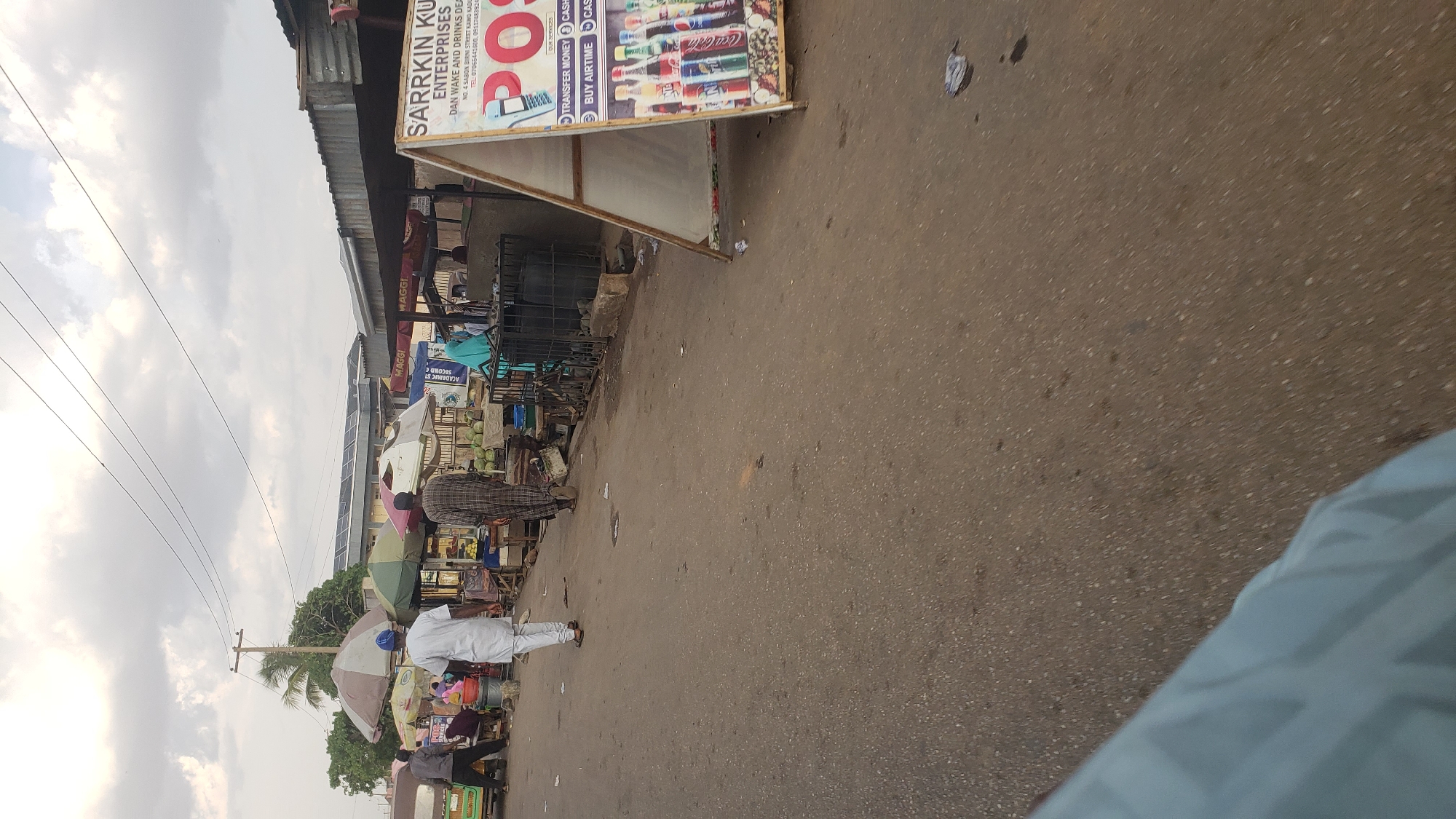 The image shows a street scene in Kawo, Nigeria. A sign for "SARRKIN KU ENTERPRISES" advertises money transfer, airtime, and cash services. Pedestrians walk along the street. Various vendors and shops line the side of the street, sheltered by awnings. The street surface is paved. Overhead electrical wires run along the street. The sky is overcast.