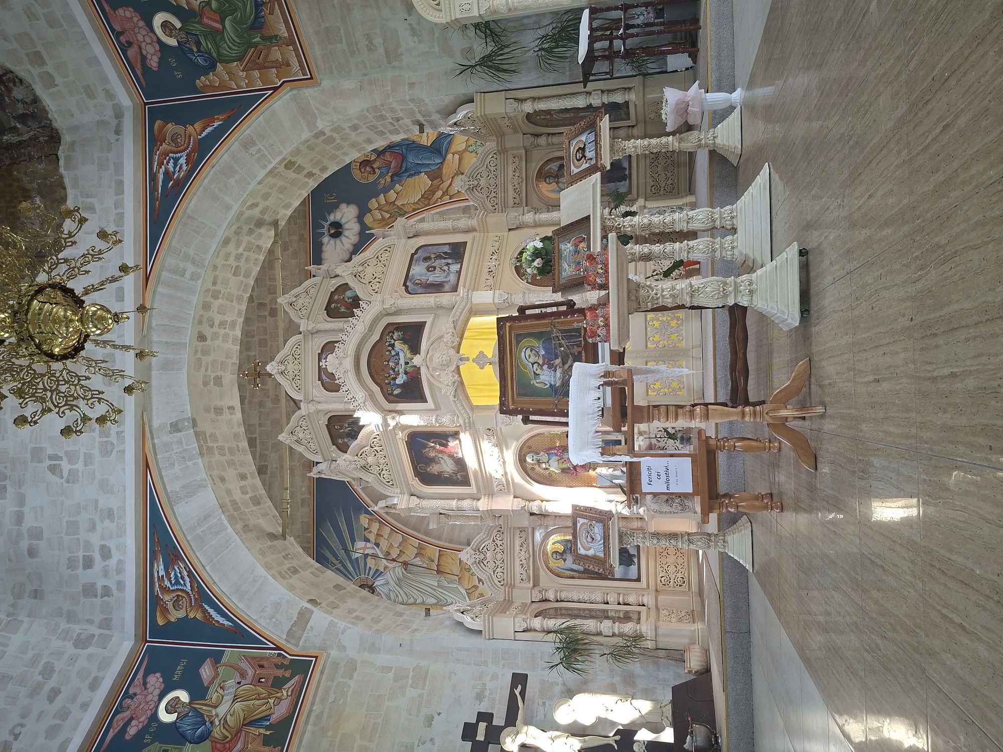 This image depicts the interior of an Orthodox church in Condrița, Moldova. The scene is dominated by an ornate iconostasis, a decorative screen with icons that separates the sanctuary from the nave. The iconostasis is intricately carved and adorned with religious imagery, including depictions of saints and biblical scenes painted in rich colors. Above the iconostasis, a fresco adorns the arched ceiling, featuring more religious artwork and architectural details.
In the foreground, the polished tiled floor reflects the ambient light. Several wooden stands are placed on the floor, holding religious icons and what appears to be a prayer book. A white cloth is draped over one of the stands. A sign with text in Romanian is also visible on one of the stands. To the left of the iconostasis, a large crucifix with a statue of Jesus is mounted on a wall, accompanied by another statue. Potted plants are positioned near the walls, adding a touch of greenery to the scene. A large, ornate chandelier hangs from the ceiling, providing light. The walls are constructed of light-colored stone blocks, consistent with traditional church architecture. The overall impression is one of solemnity and religious devotion.