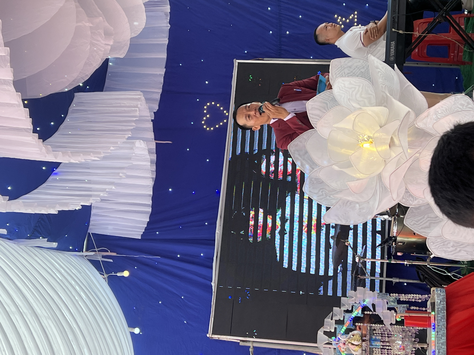 The image shows an indoor event in Tay Son Commune, Vietnam. A man in a red blazer is speaking into a microphone in front of a large video screen, while another man wearing a white polo shirt is seen in the background. There are several large white flower decorations, and draped white fabric hangs from the ceiling. A Yamaha keyboard is visible behind the man in the white polo shirt. Other decorations include a string of lights that appear in the foreground. The backdrop is a dark blue curtain.