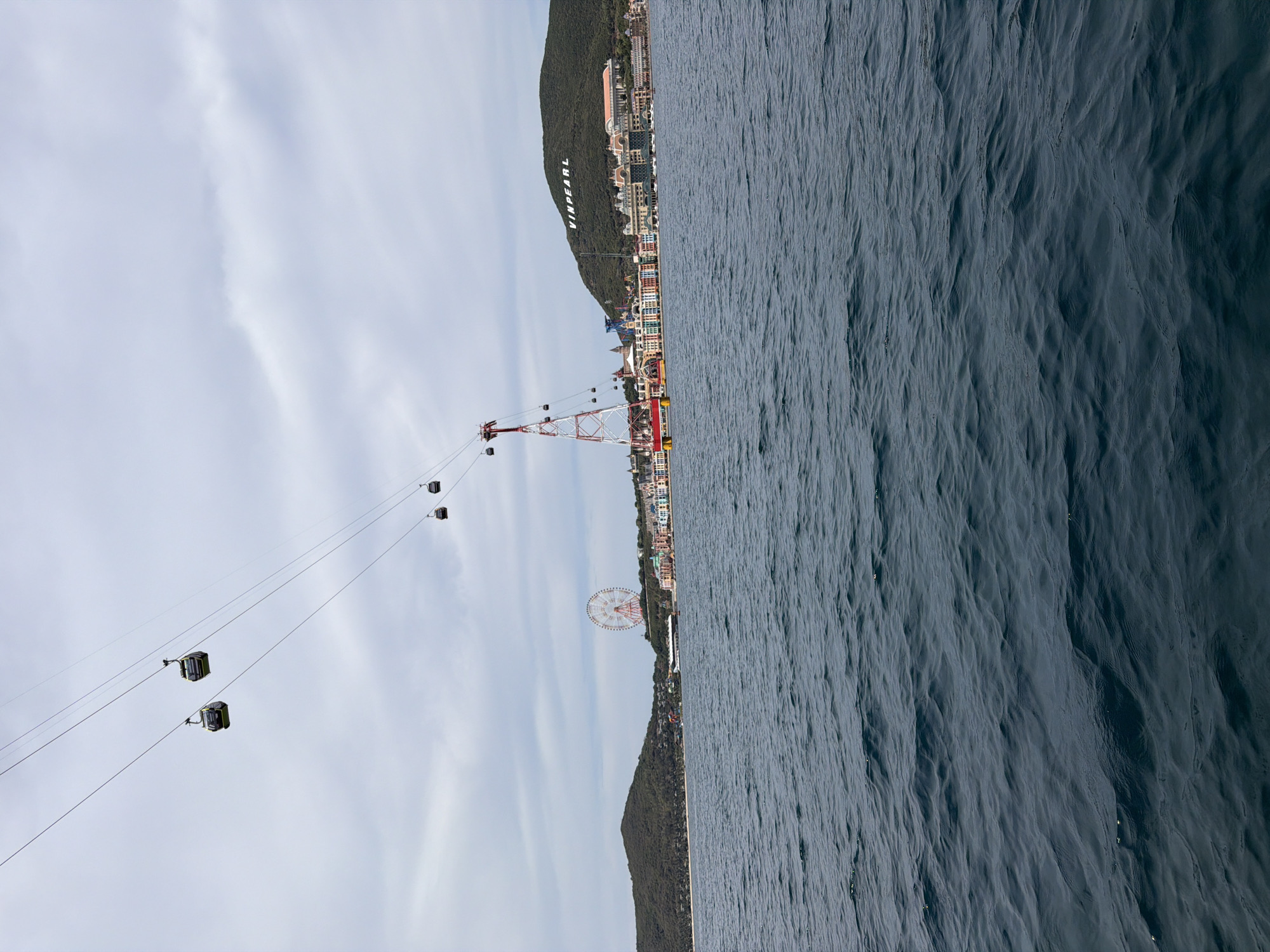 This image shows a coastal scene in Nha Trang, Vietnam. The focus is on a cable car system stretching from the shore out over the water. The cables are visible, with several enclosed cable cars suspended along the line.
A prominent structure is a tall, red and white cable car support tower located near the coastline. In the background, there is a shoreline with buildings and structures, including what appears to be a Ferris wheel and "VINPEARL" written on the mountain. To the right of the shoreline, there is water.
The sky above is overcast with clouds.