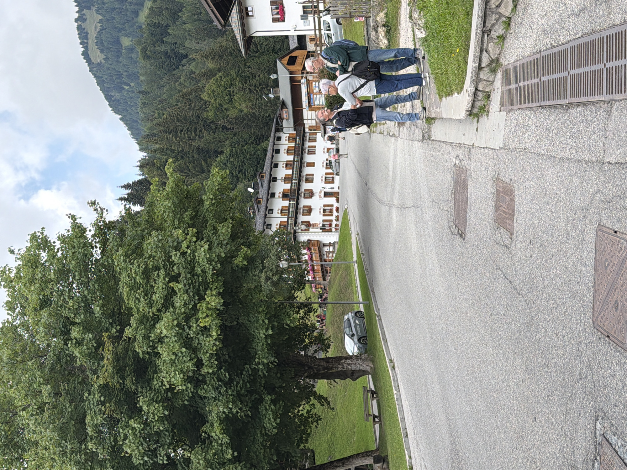 The image depicts a street scene in Sauris di Sopra / Plozn / Sauris Disore, Italy. In the foreground, three individuals, two men and a woman, are engaged in conversation near a street drain and a stone-lined garden bed. The man on the left, wearing a black jacket and blue jeans, faces the other two. The woman, in a light-colored top and blue jeans, stands between the two men, her hands touching the man to her left. The man on the right, wearing a green jacket and blue jeans, looks towards the man on the left.
Further down the street, several buildings line the left side of the road. The most prominent is a multi-story building with white walls, dark shutters, and balconies, suggesting a traditional alpine architectural style. Across the street, a large, leafy tree dominates the left side of the frame, with a wooden bench beneath it. A small, gray car is parked under the tree. In the background, a grassy slope rises behind the buildings, leading to a dense forest of evergreen trees that covers a mountain. The sky above is partly cloudy. The overall impression is a quiet, scenic setting in a mountainous Italian village.