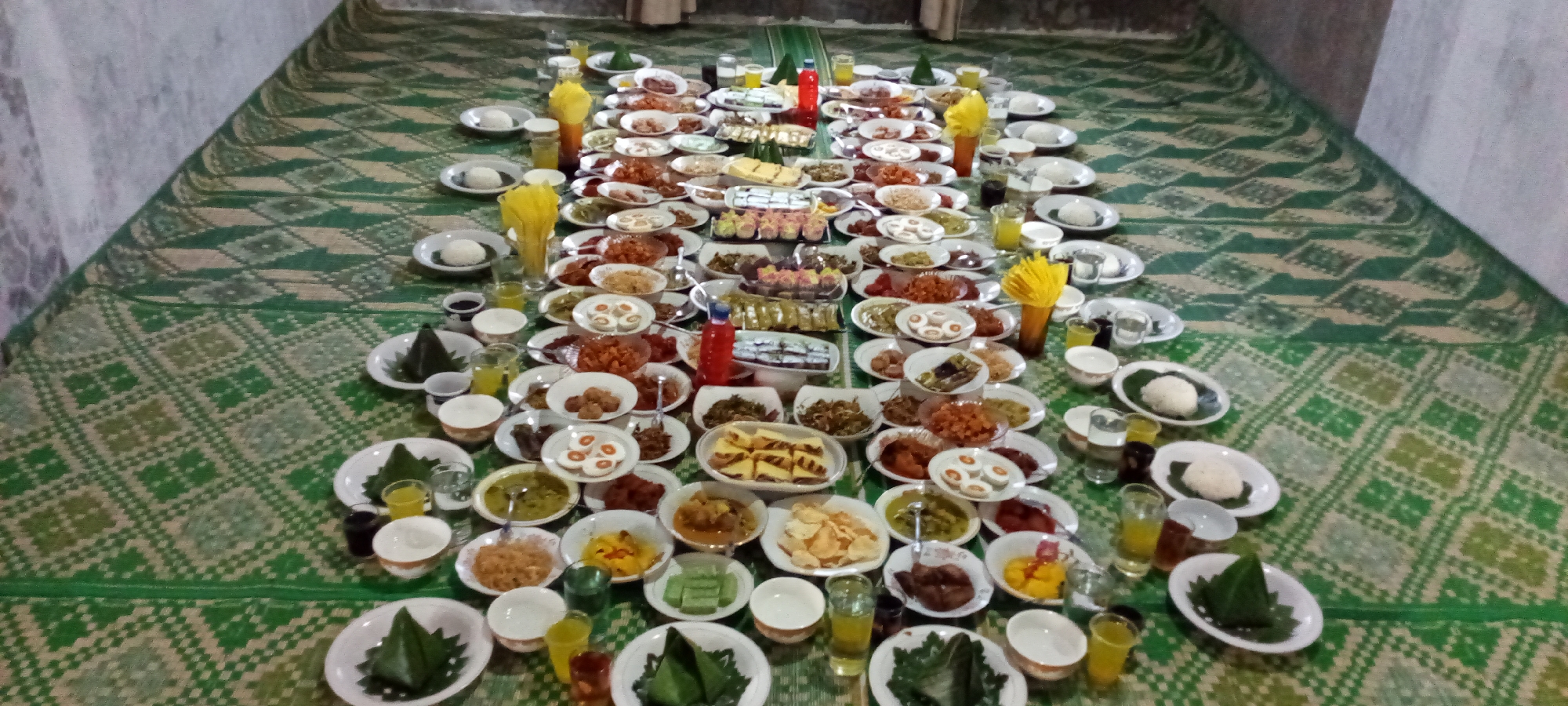 This image captures a wide-angle view of a traditional communal meal spread laid out on a green and beige patterned mat, likely in a room in Lhokseumawe, Indonesia. The mat features repetitive geometric and floral motifs. Numerous white ceramic plates and bowls are meticulously arranged in long, parallel rows extending from the foreground towards the distant background, covering the majority of the floor space.
The spread consists of a vast assortment of Indonesian dishes. Many small white plates contain individual portions of white steamed rice, often accompanied by small clear glasses filled with a light yellow liquid (likely juice) or water, and smaller white bowls. Interspersed between these individual settings are larger white plates and rectangular trays holding a variety of main courses, side dishes, and desserts. Observable dishes include items with reddish-orange sauces, sliced hard-boiled eggs, various vegetable preparations, fried items (like crackers or chips), what appear to be meatballs, and some items wrapped in dark green banana leaves, possibly traditional snacks or rice portions. Several decorative elements, such as tall clear vessels containing scrunched yellow material (likely napkins or fabric), are placed at intervals along the rows.
The background reveals plain, light-colored walls with a slightly rough texture. Hints of furniture or draped fabric are visible in the distant background, suggesting an indoor setting. No individuals are present in the frame, but the extensive arrangement of food indicates preparation for a large gathering or an ongoing event where people are momentarily out of view. The lighting appears to be artificial, evenly illuminating the scene.