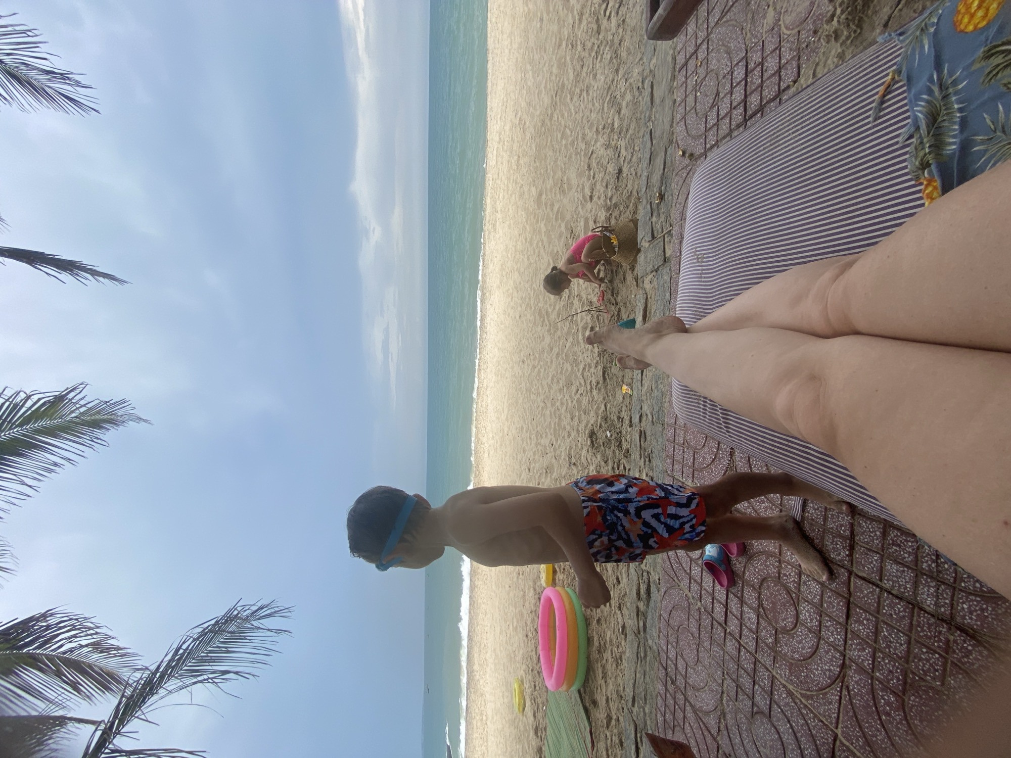 A young boy wearing blue goggles and colorful swim trunks stands on a sandy beach in Phu Thuy Ward, Vietnam. He is looking out towards the ocean, with waves gently lapping at the shore. The sky is a bright blue with some scattered clouds, suggesting a pleasant day. Palm fronds frame the top and left side of the image, indicating a tropical or subtropical location.
In the background, a young child in a pink swimsuit is playing in the sand, near a straw basket. A multi-colored inflatable ring lies on the sand near the boy. The foreground shows a person's legs and feet, resting on a striped lounge chair with a tiled pathway beside it. The overall atmosphere is relaxed and summery, with a focus on a family enjoying a day at the beach. The time of day appears to be daytime, possibly late morning or early afternoon, given the brightness of the sky. No visible text is present in the image.